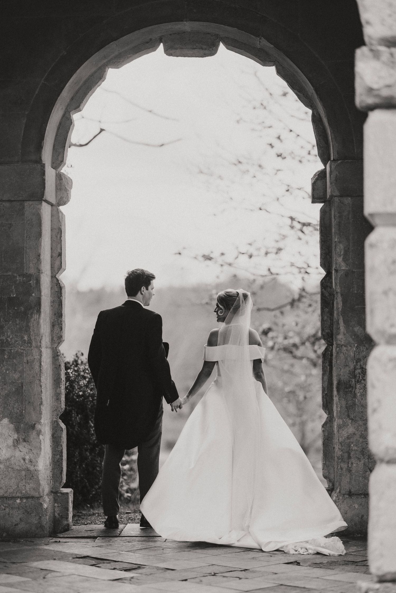 A black and white wedding photo sequence showing a couple walking through stone archways in elegant formal attire.