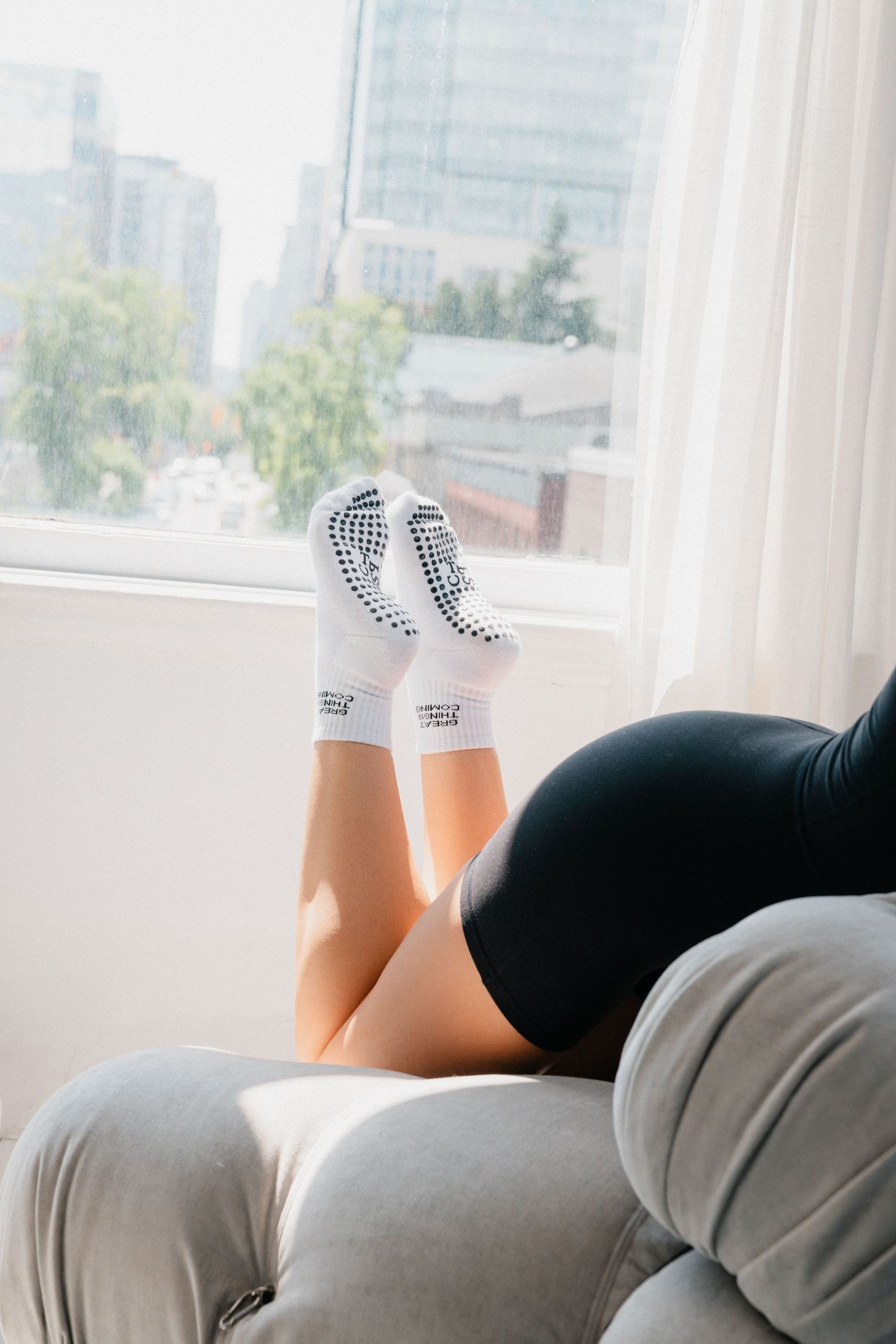 Person wearing black shorts and checkered socks relaxes with legs up against white cushions by a bright window.