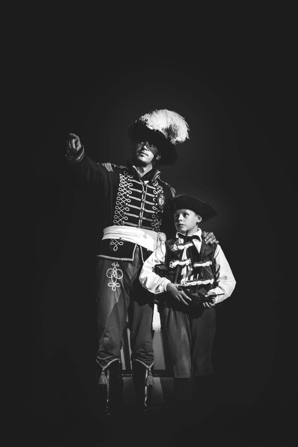 Black and white photo of two performers in elaborate historical costumes on a dark stage.