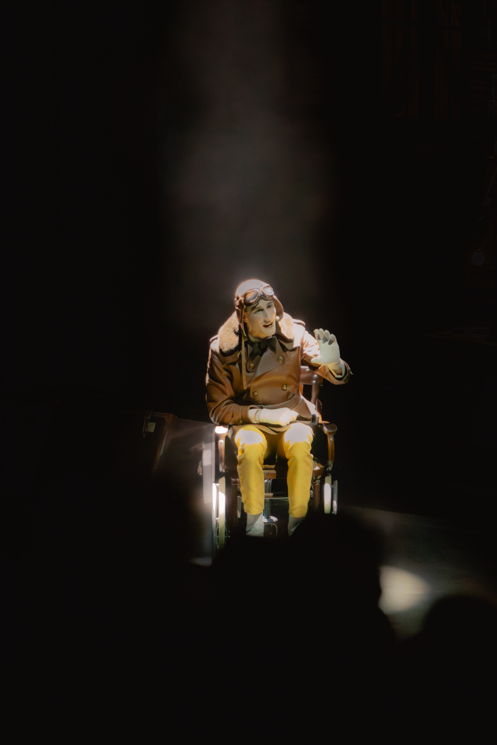 Performer in vintage aviator costume seated in wheelchair under dramatic spotlight on dark stage.