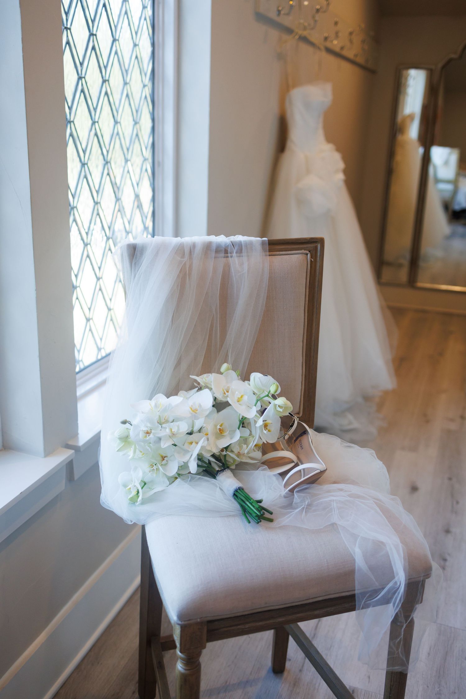 A white wedding bouquet and veil rest on an upholstered chair with a bridal gown in the background in the bridal suite at Bella Cavalli