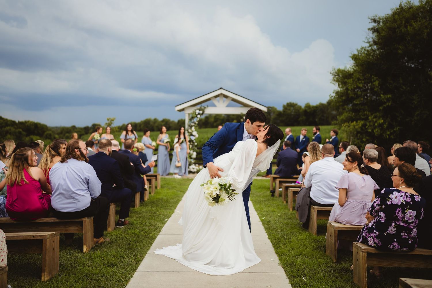 A romantic outdoor wedding ceremony with guests seated on wooden benches against a scenic countryside backdrop.