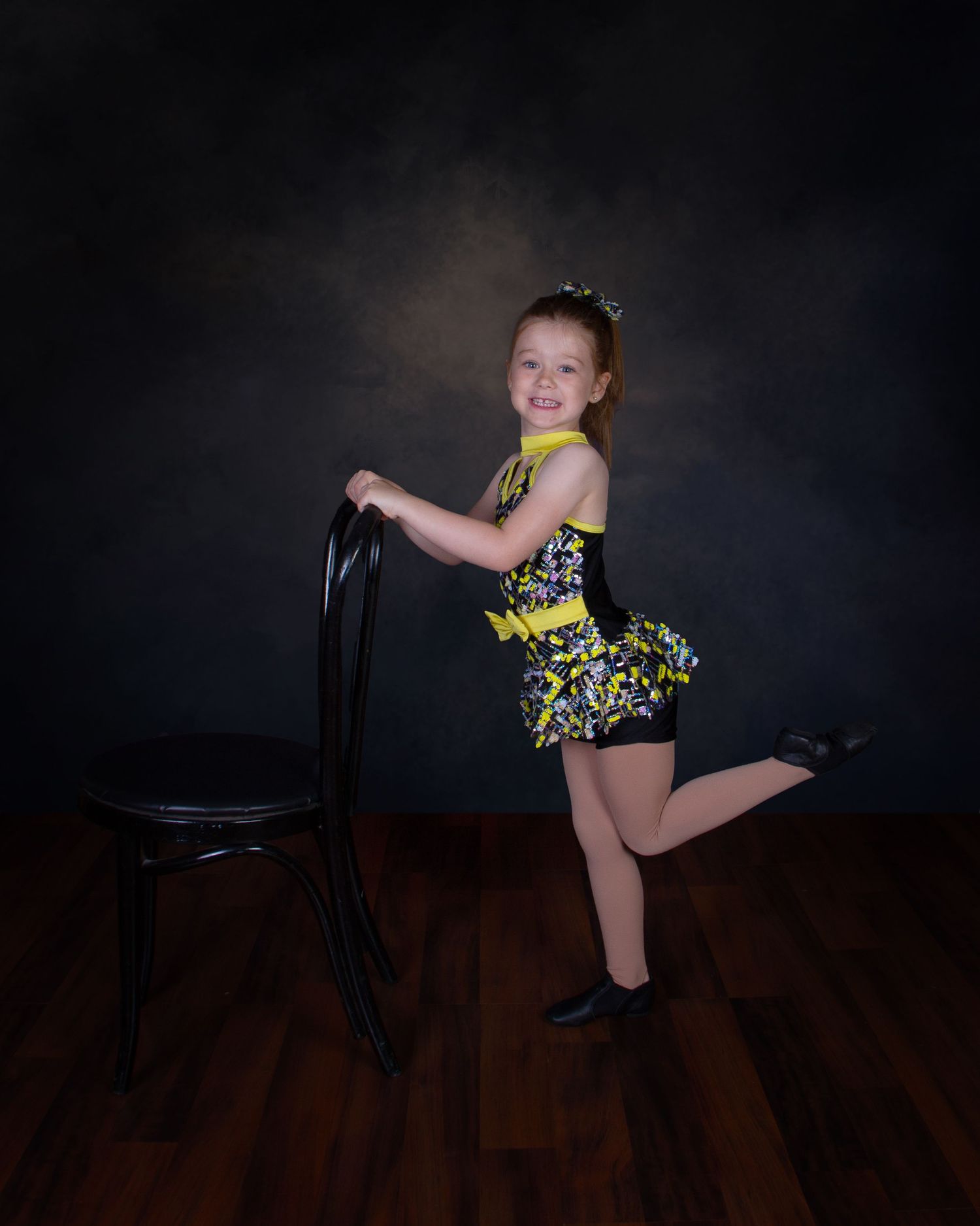 Young dancer in pink tutu and cat ears poses with arms outstretched on a dark wooden stage floor.