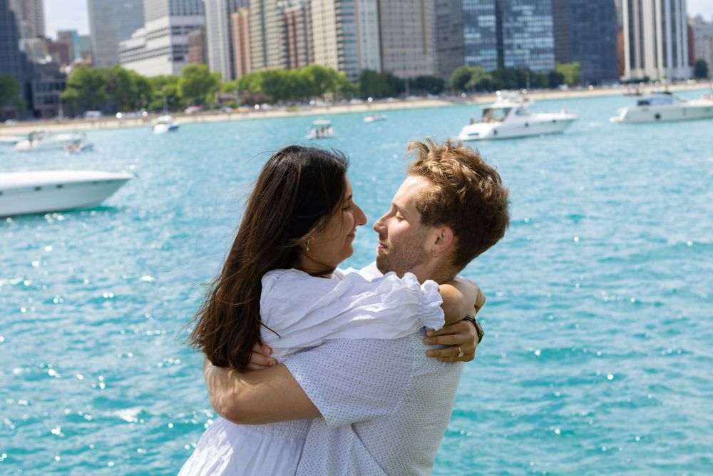 Couple in white embracing by turquoise lake with city skyline and boats in background.