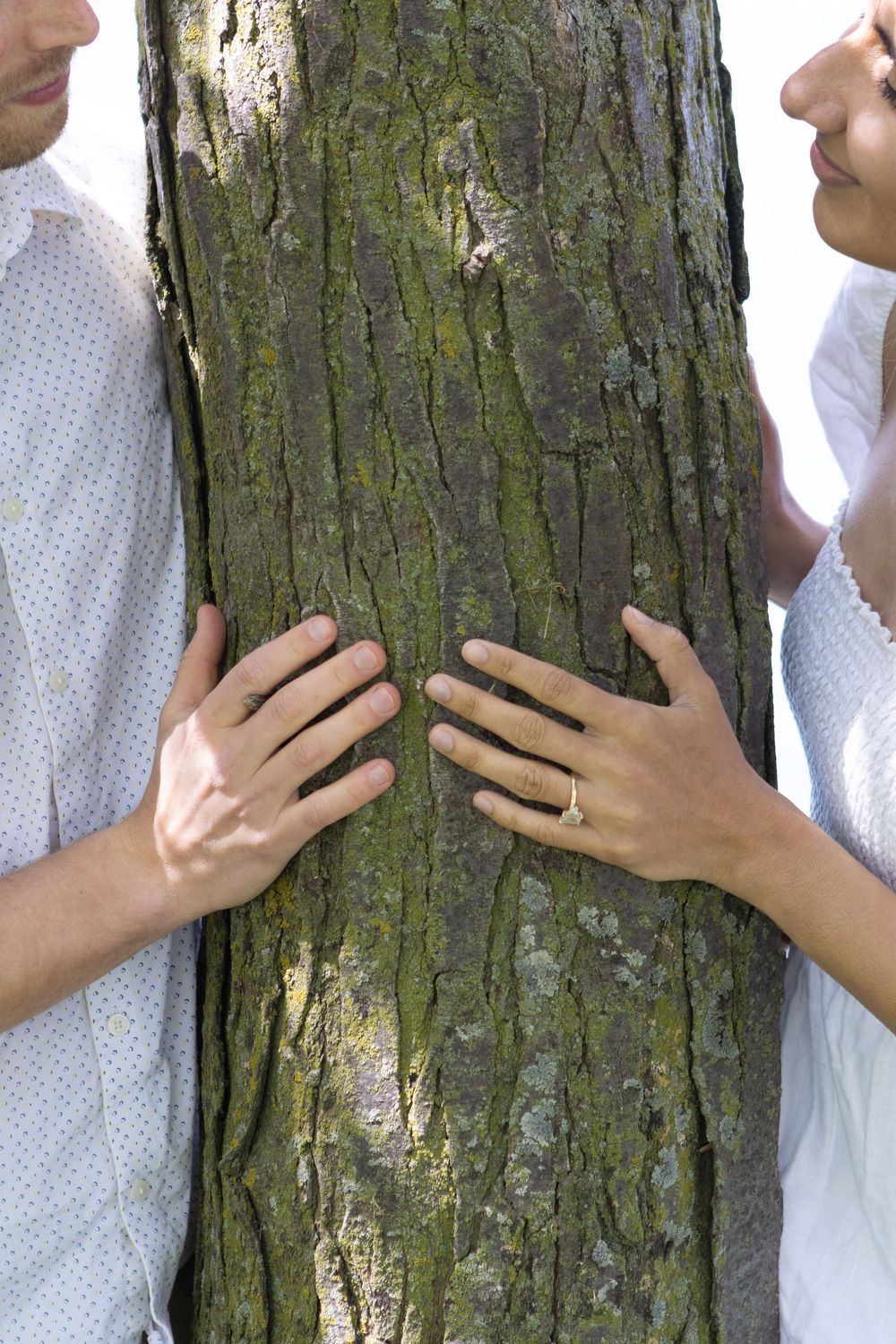 Couple embracing a large tree trunk outdoors, with a diamond engagement ring visible on the woman's hand.