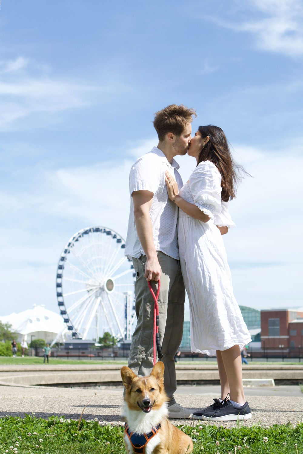 Couple kissing outdoors with a Corgi dog on a red leash, ferris wheel in background on a sunny day.