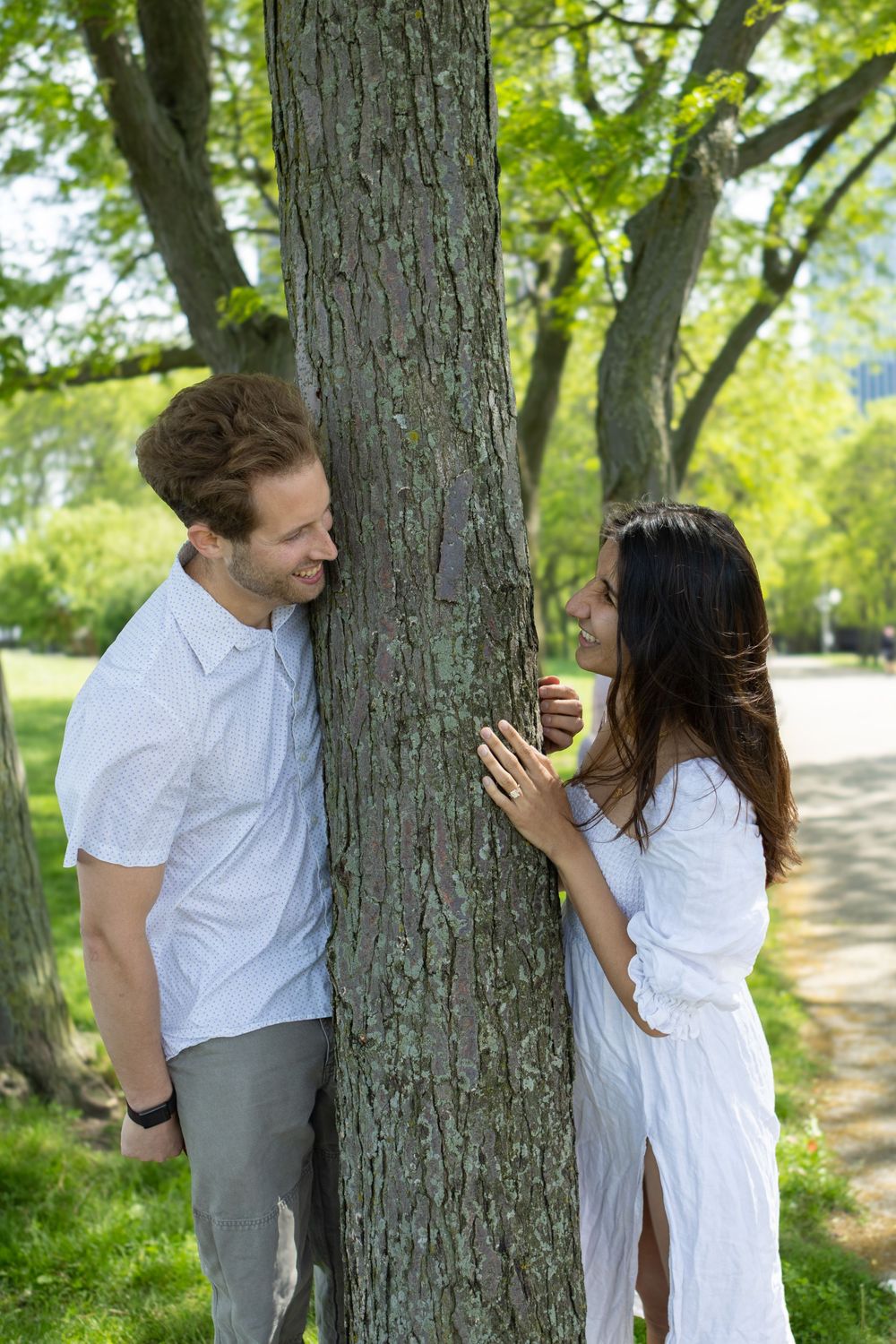 Smiling couple playing peek-a-boo around a tree trunk in a lush green park.