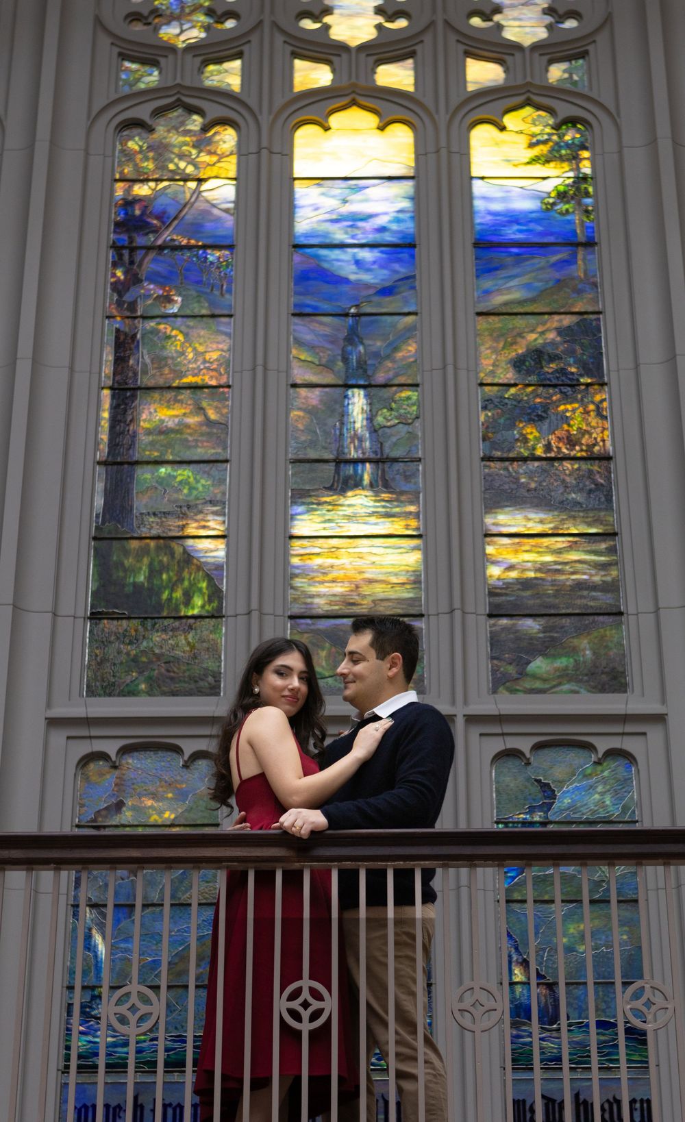 A couple shares a romantic moment in front of ornate stained glass windows in a Gothic church setting.