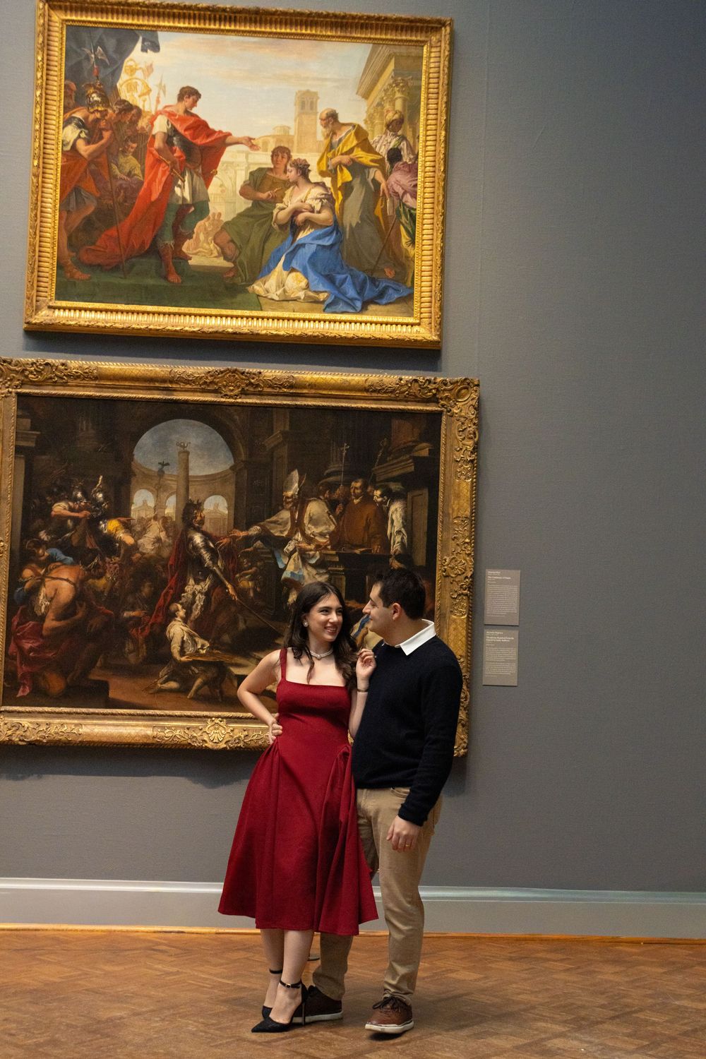 A couple in formal attire dances together in an art museum gallery surrounded by classical paintings in ornate frames.
