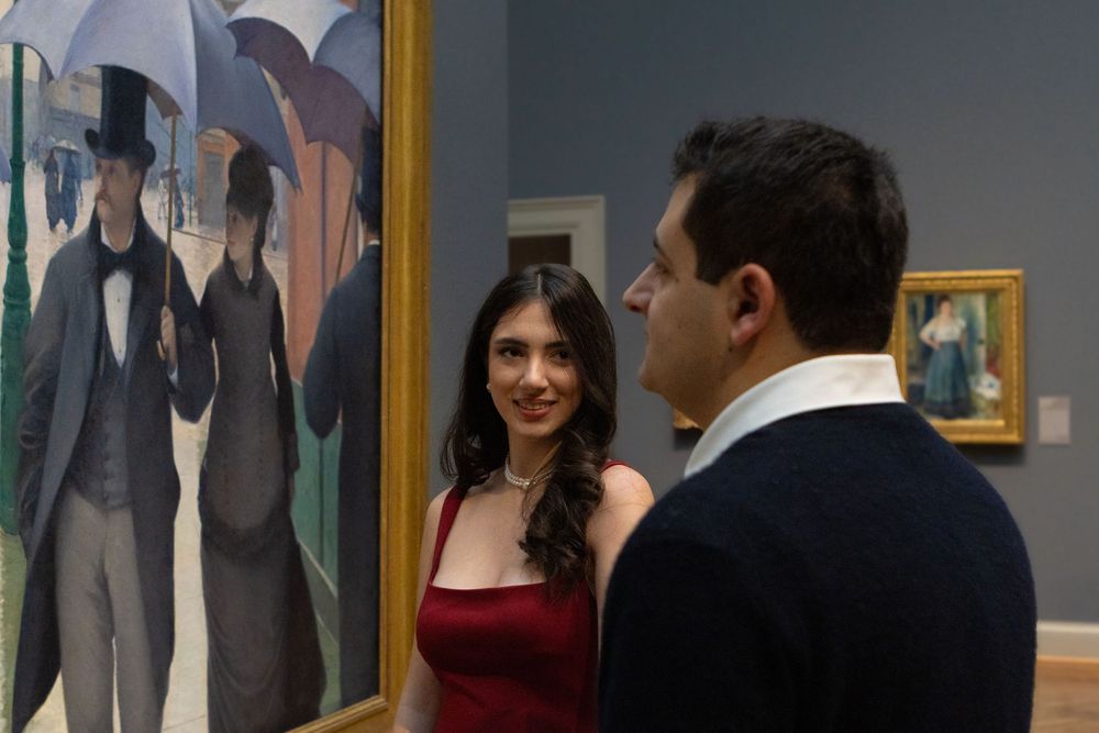 Two people having a conversation in an art gallery with a painting of figures with umbrellas visible in the background.