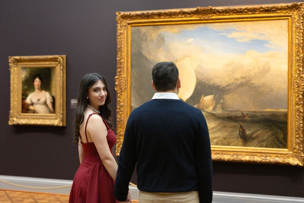 Museum visitors admire classical paintings in ornate gold frames hanging on dark walls of an art gallery.