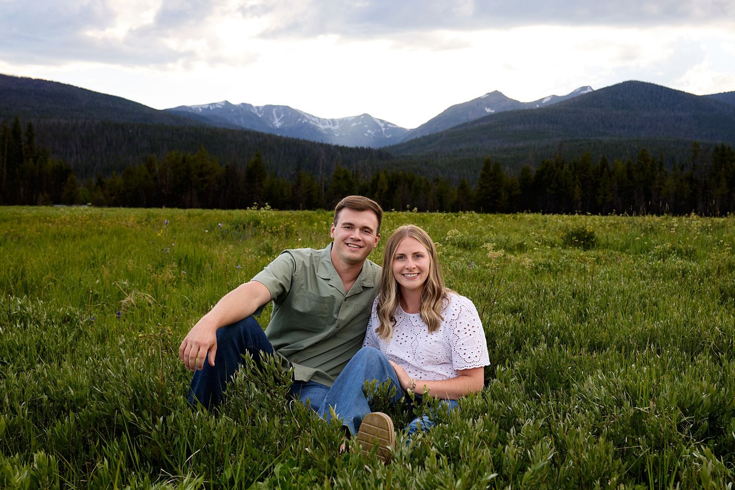 A couple sits together in a green meadow with snow-capped mountain peaks and forest visible in the background.