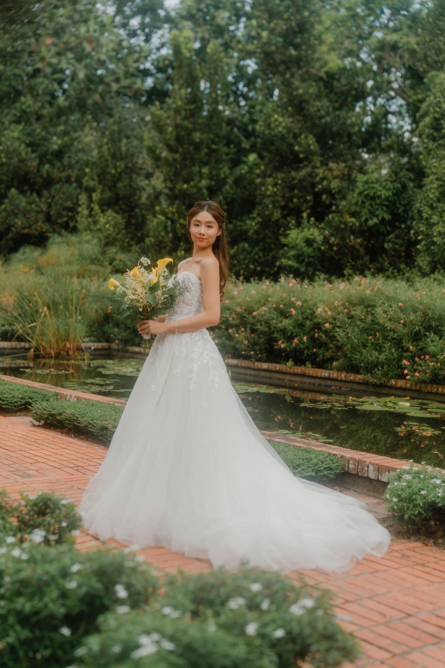 A bride in a flowing white tulle ballgown poses in a garden setting with brick pathways and lush greenery.