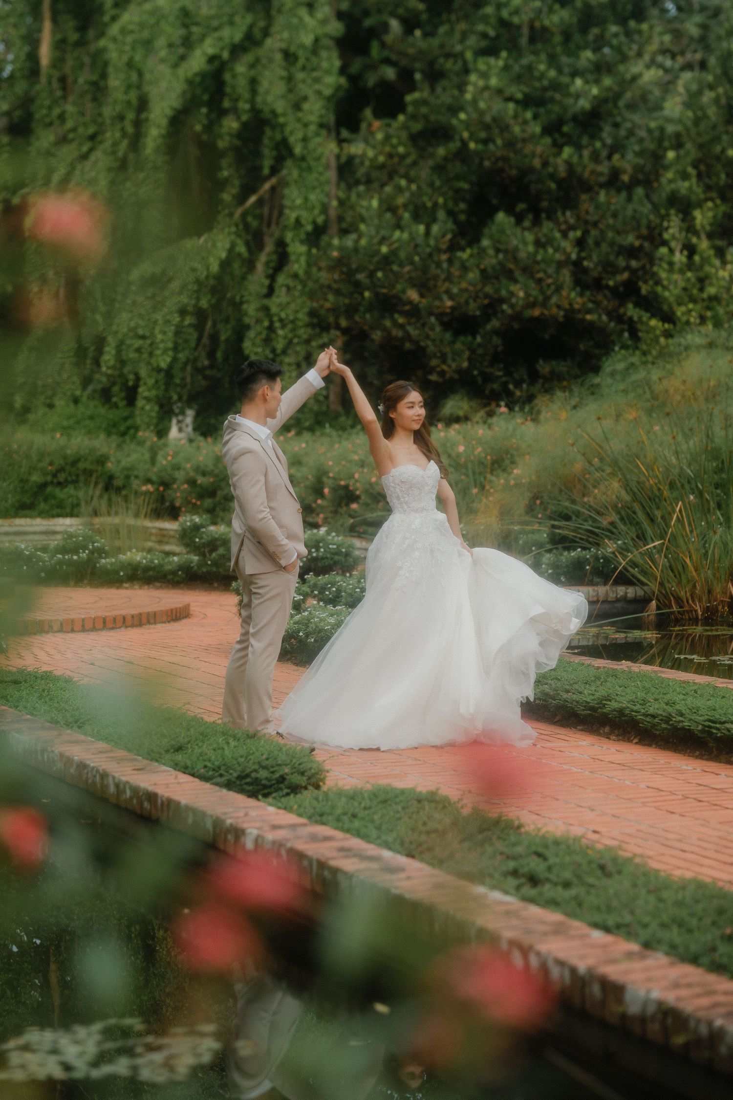 A couple in wedding attire dancing elegantly on a brick pathway surrounded by lush garden greenery.