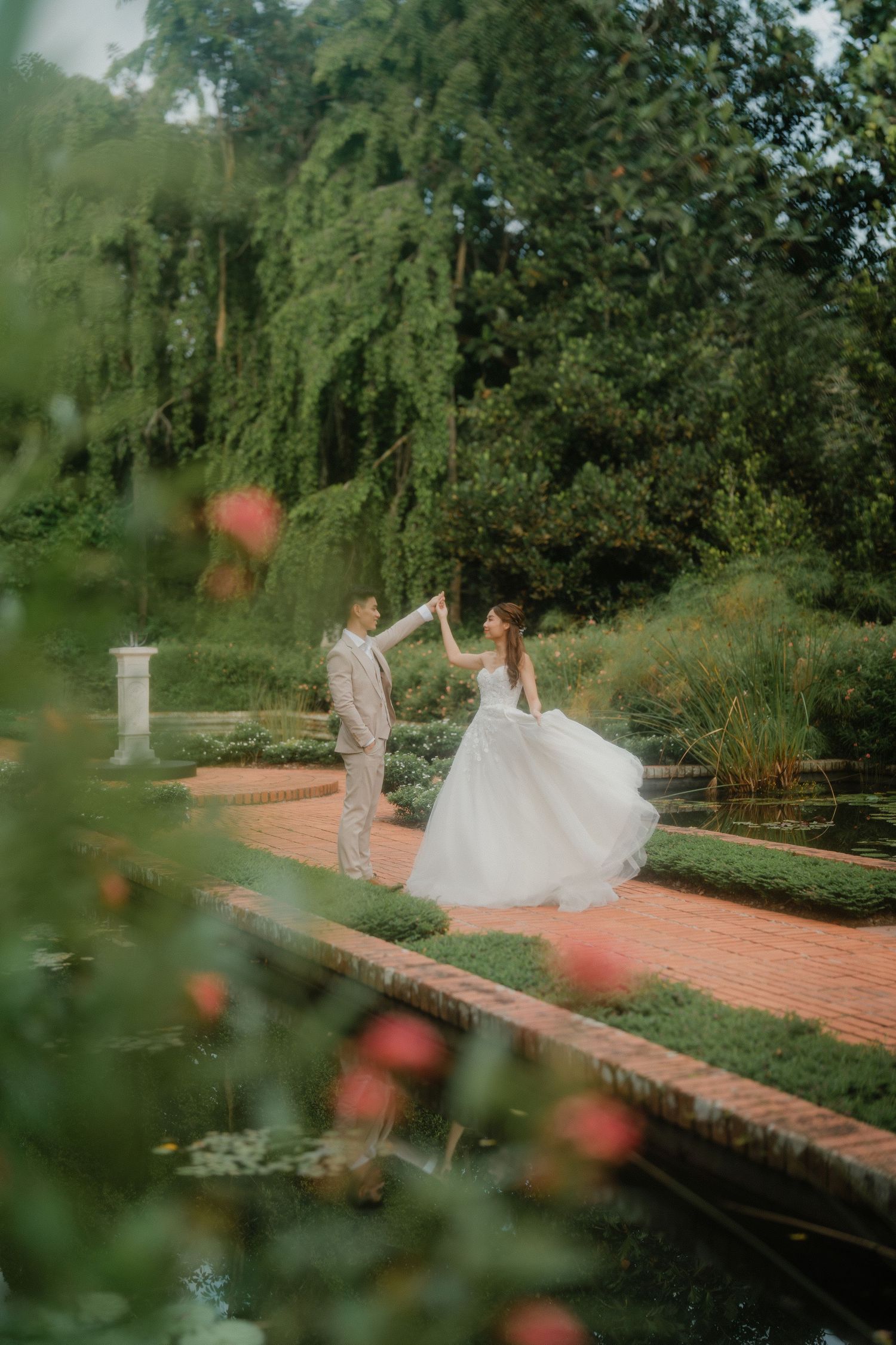 A couple in wedding attire dance together on a brick pathway in a lush garden setting at sunset.