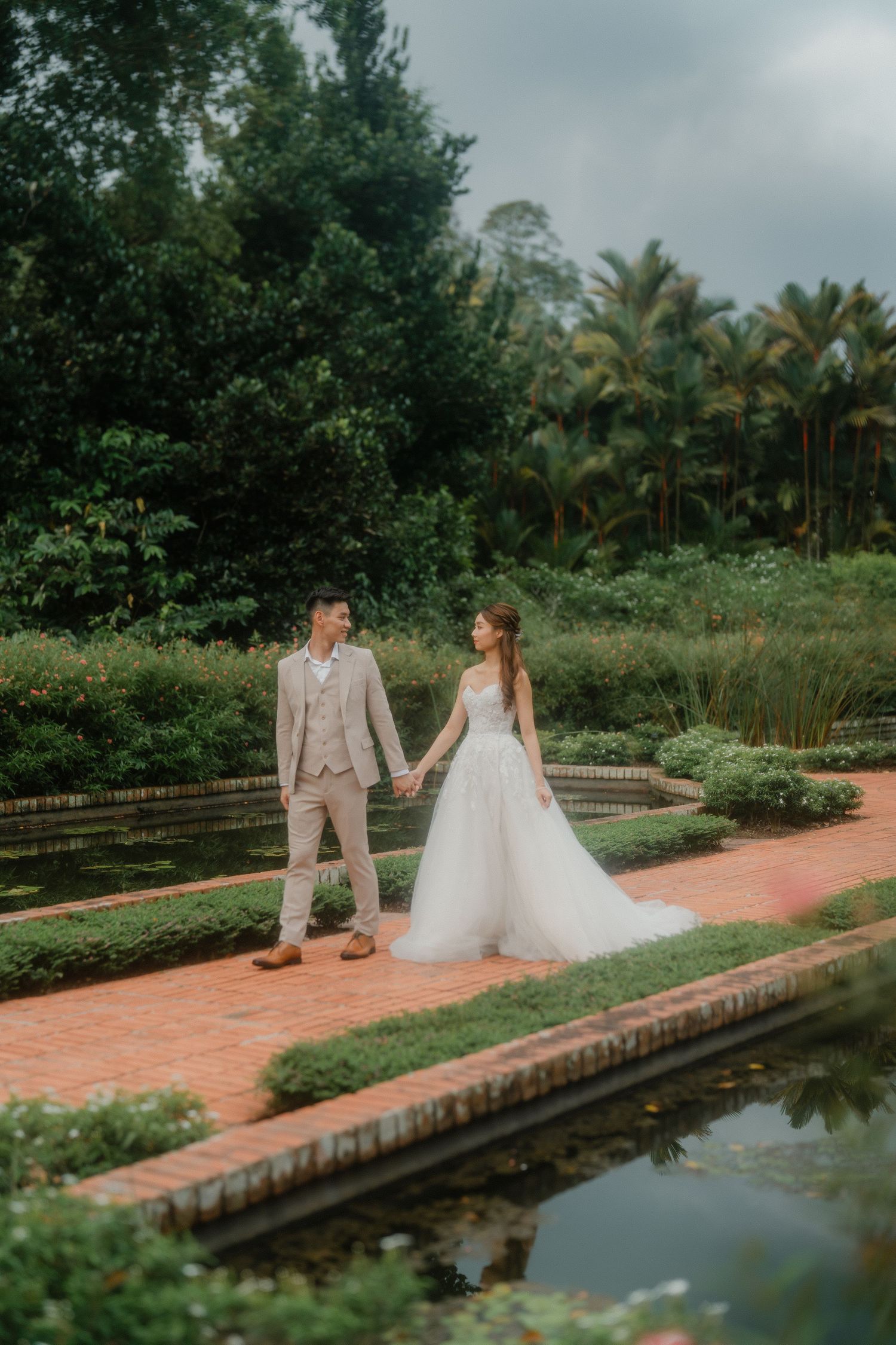 Couple in wedding attire walking together on a brick pathway beside a reflecting pond in a lush tropical garden setting.