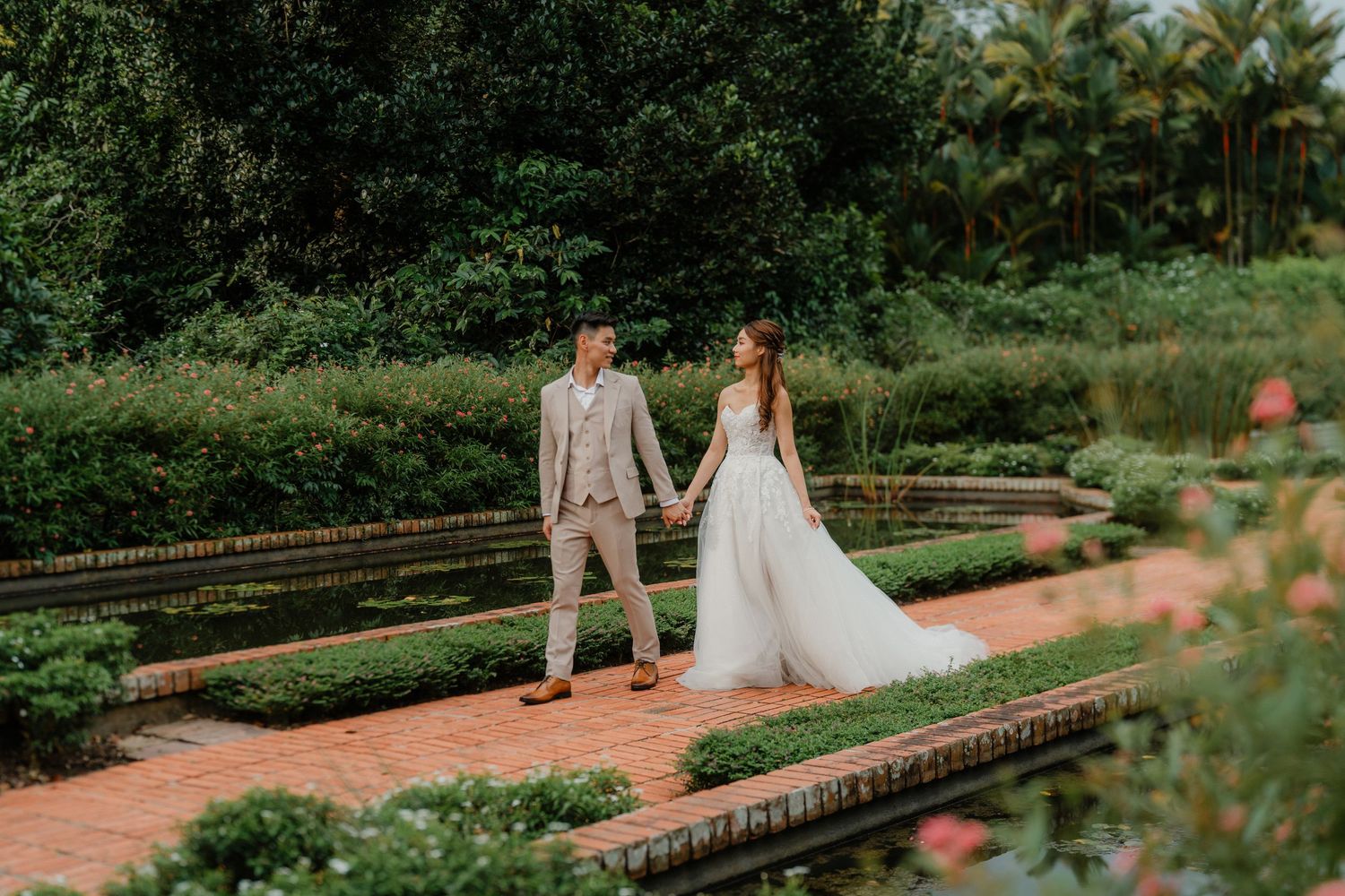 A couple strolls through a formal garden with brick pathways and manicured hedges during an outdoor wedding photoshoot.