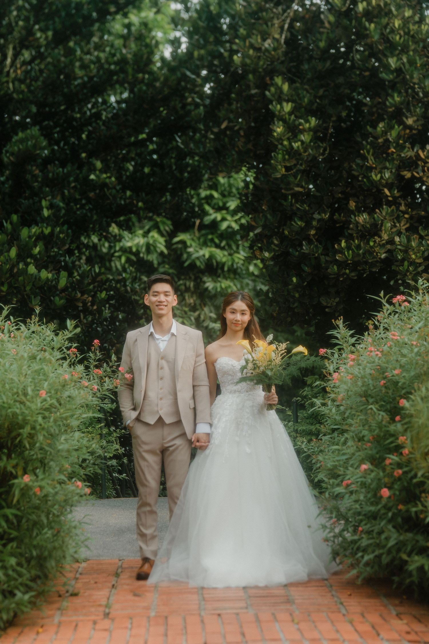 A bride and groom walk together on a brick path surrounded by lush green garden foliage on their wedding day.