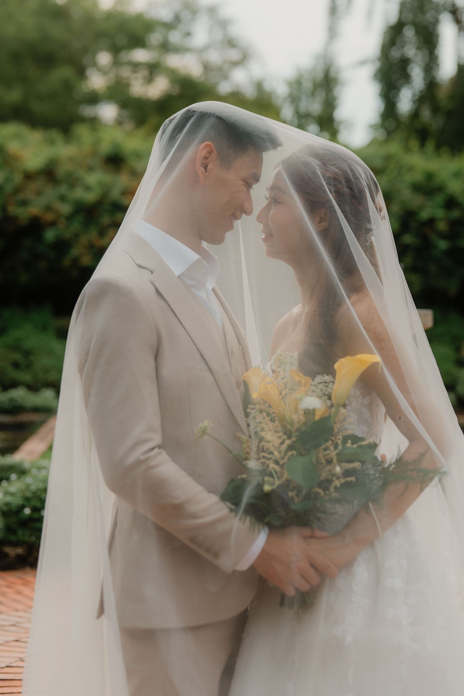 A romantic wedding moment captured outside with a couple embracing under a flowing white veil and yellow bouquet.