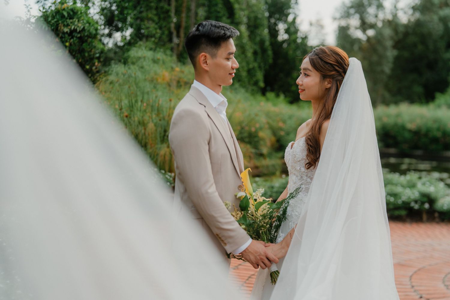 A couple in wedding attire holds hands in a lush garden setting with green foliage and trees in the background.