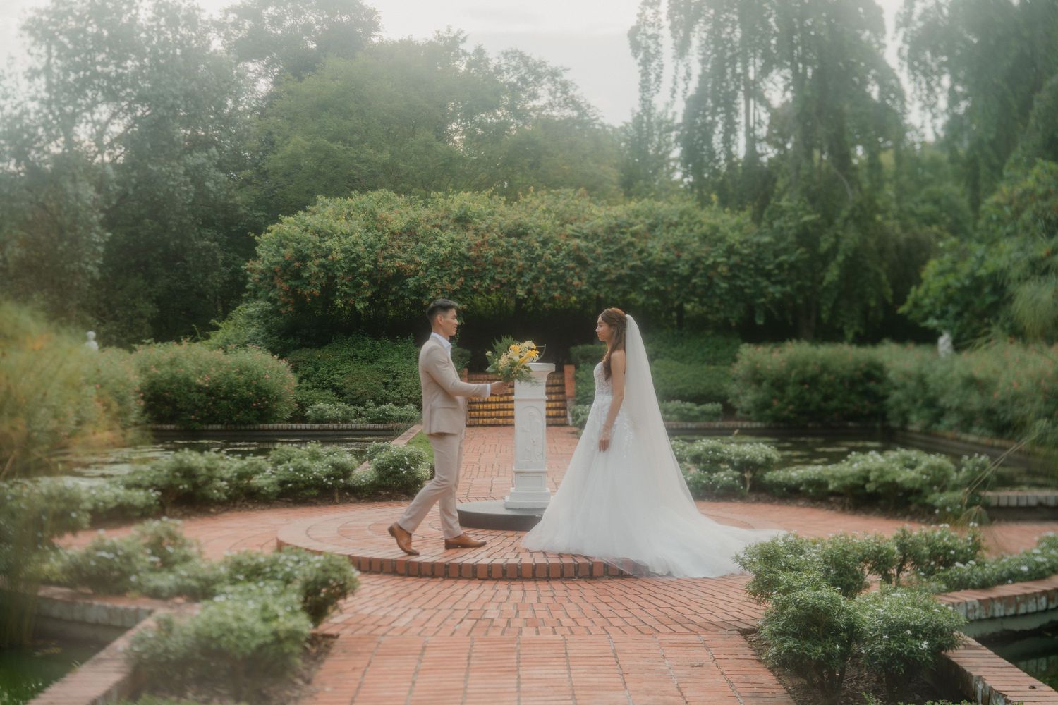 A wedding couple stands in a lush garden setting surrounded by greenery and flowering plants on their special day.