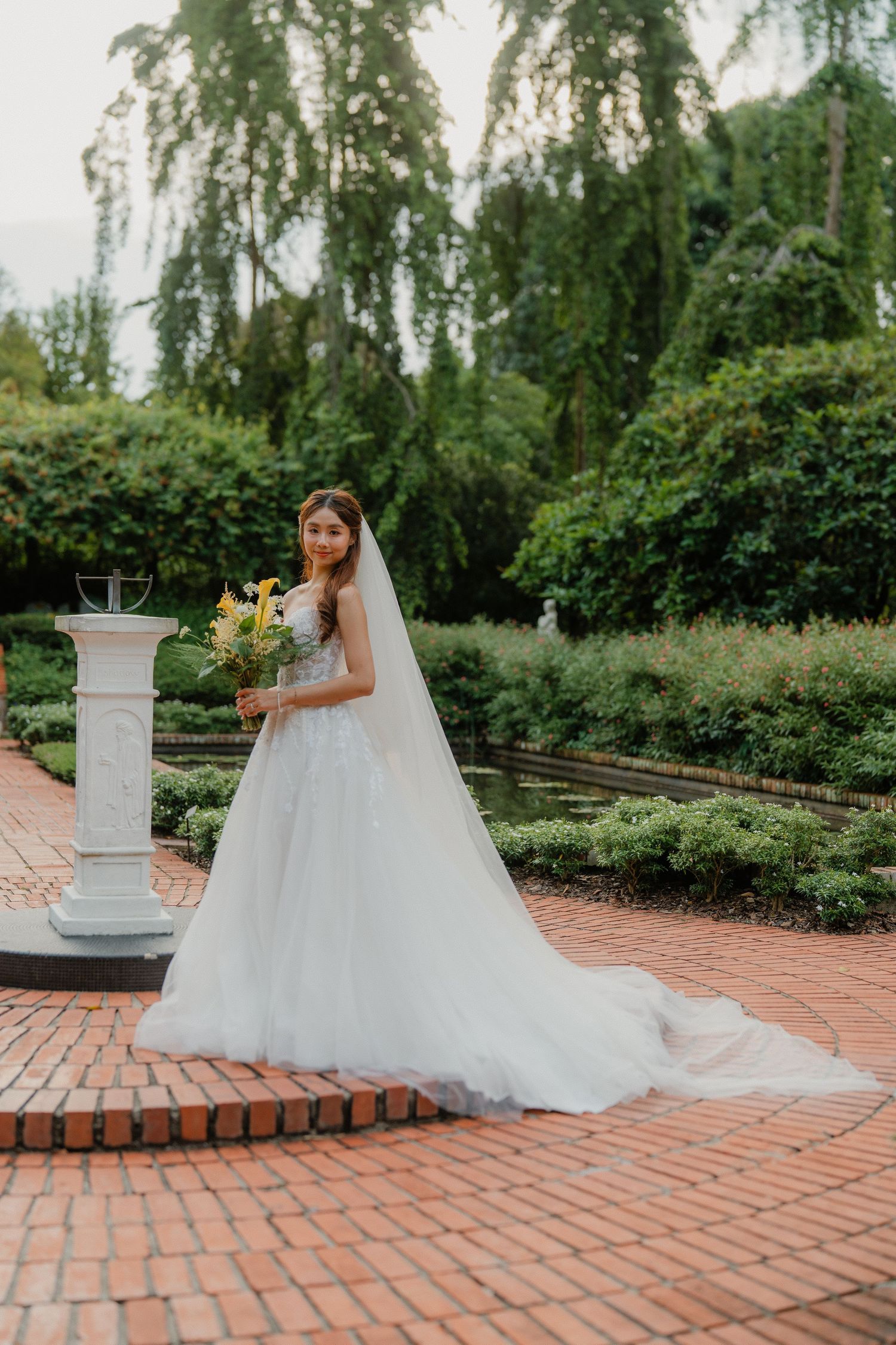 A bride in a flowing white ballgown poses gracefully on a brick pathway surrounded by lush garden greenery.