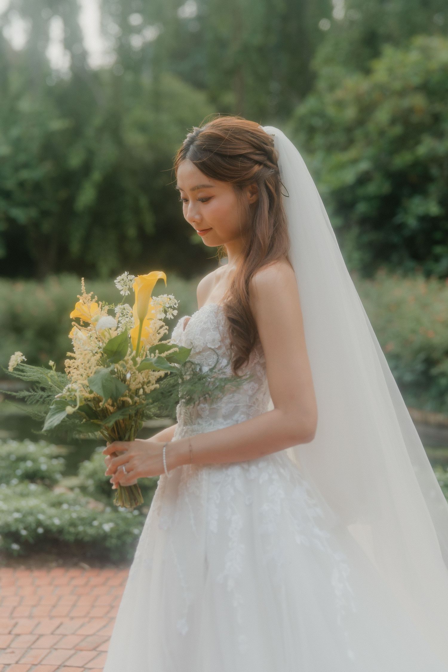 A bride in a white lace gown and long veil holds a yellow floral bouquet against a garden backdrop.