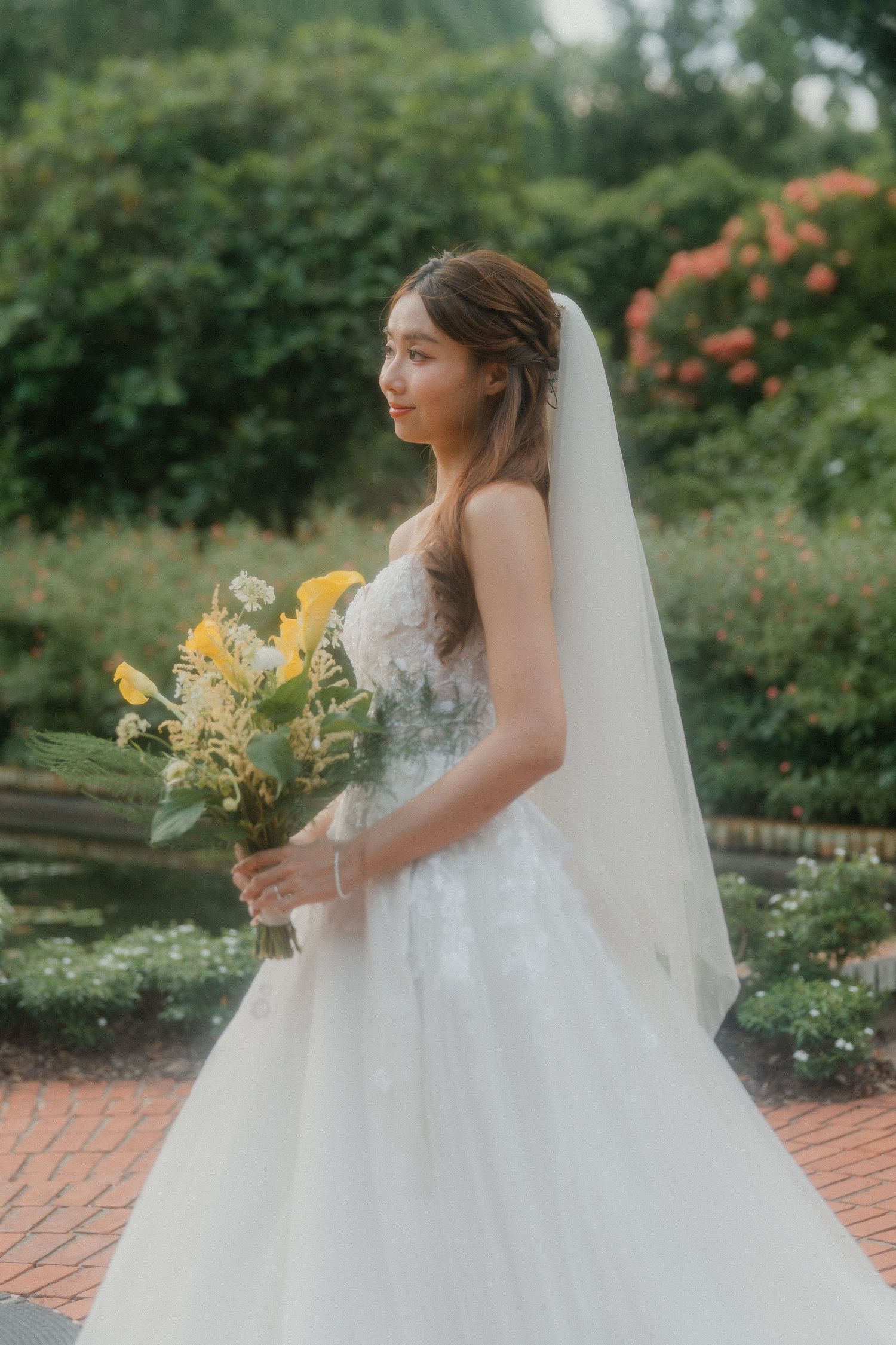 A bride in a white strapless wedding gown holds a yellow bouquet while standing in a lush garden setting.