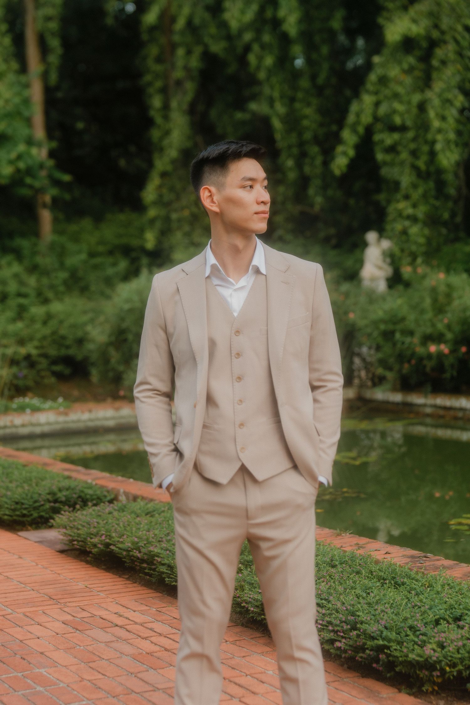 A person poses in a beige three-piece suit on a brick path surrounded by lush garden landscaping.