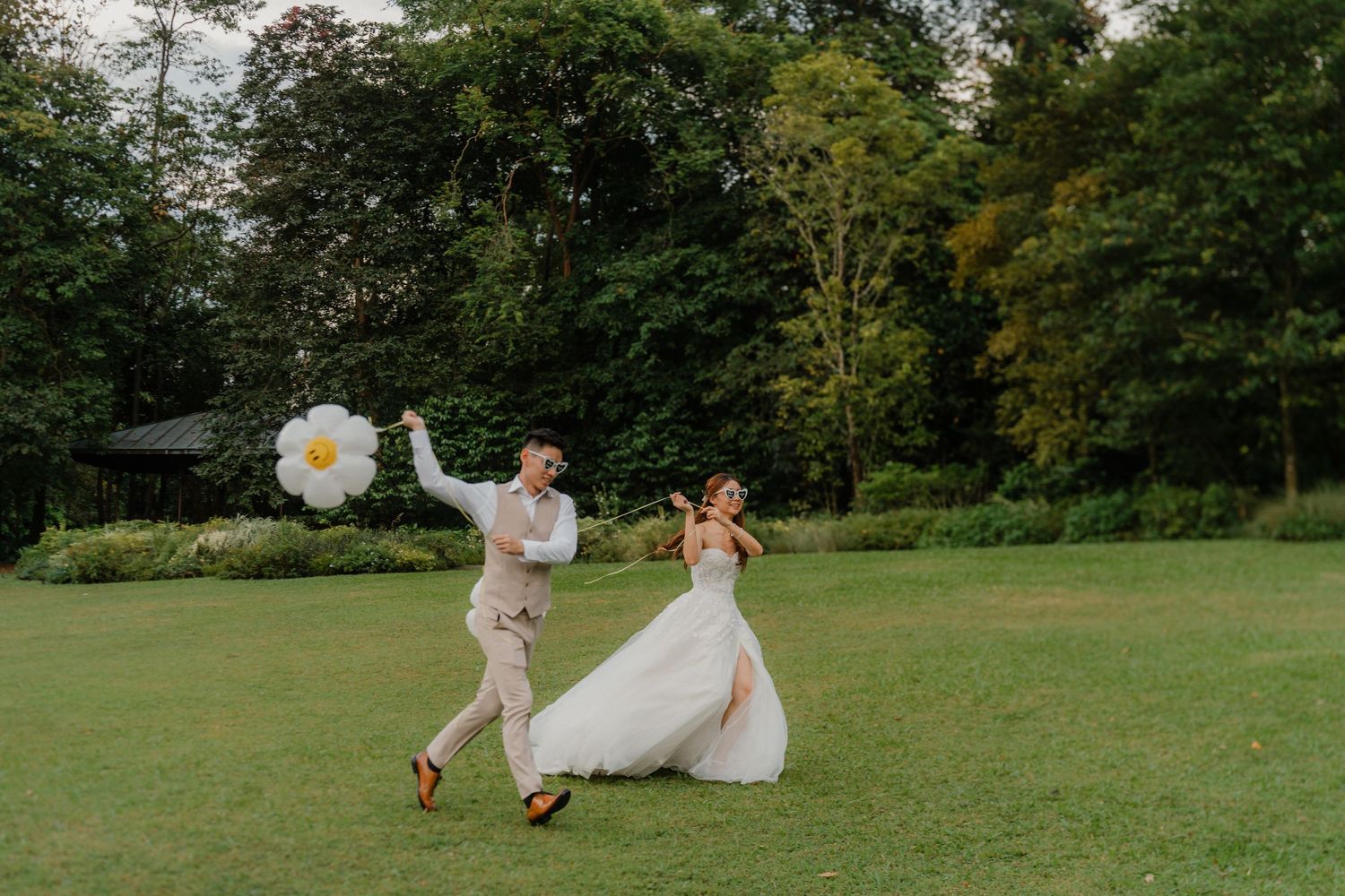 A couple walks across a lush green field as sunlight filters through trees in a romantic outdoor setting.