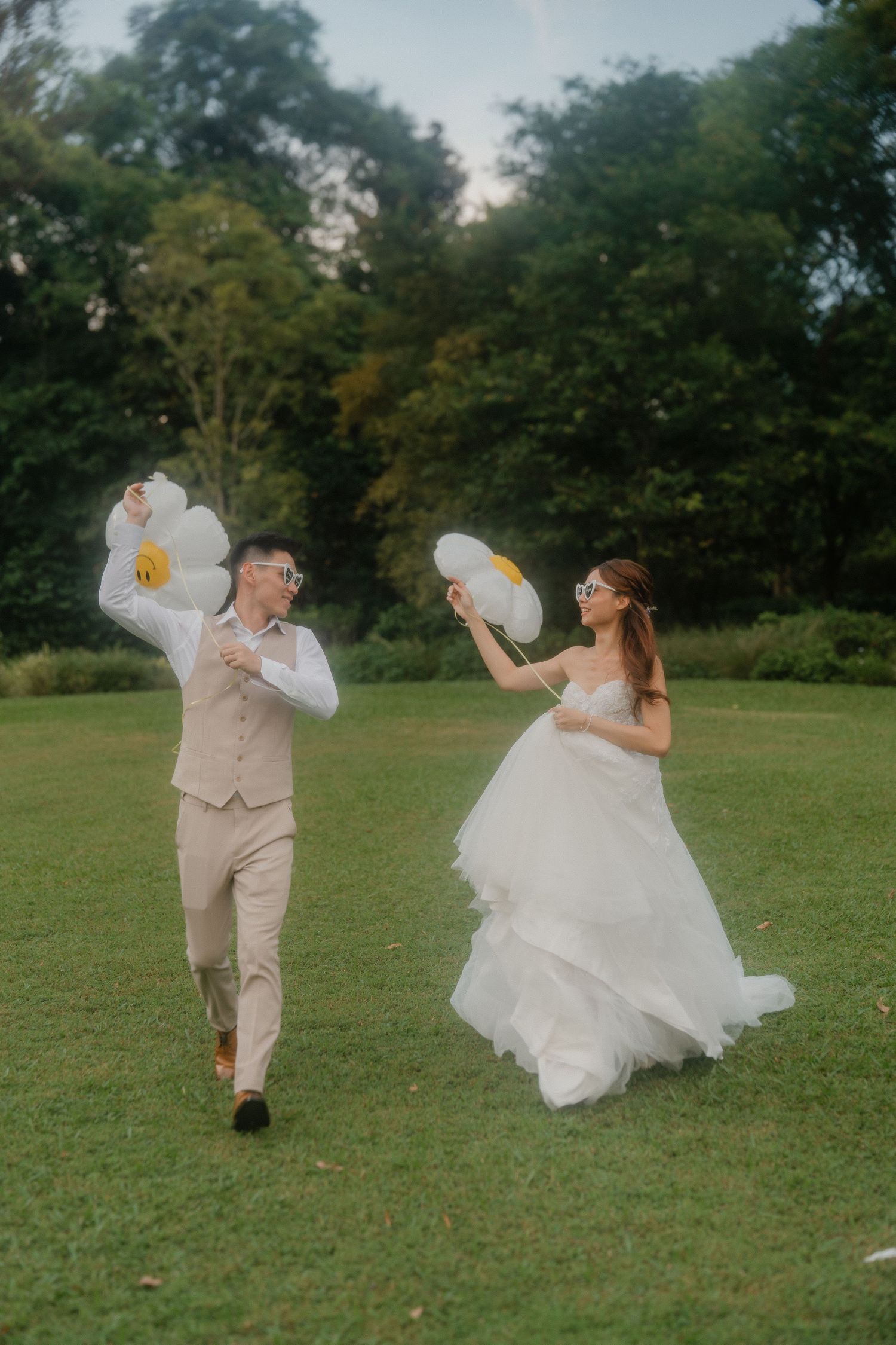 A playful wedding couple chases each other with white balloons on a grassy lawn surrounded by trees.