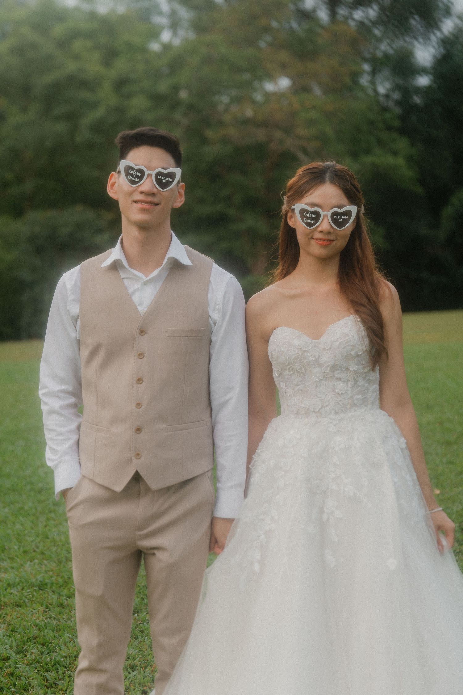 A stylish wedding couple wearing matching sunglasses poses on a grassy lawn in beige and white attire.