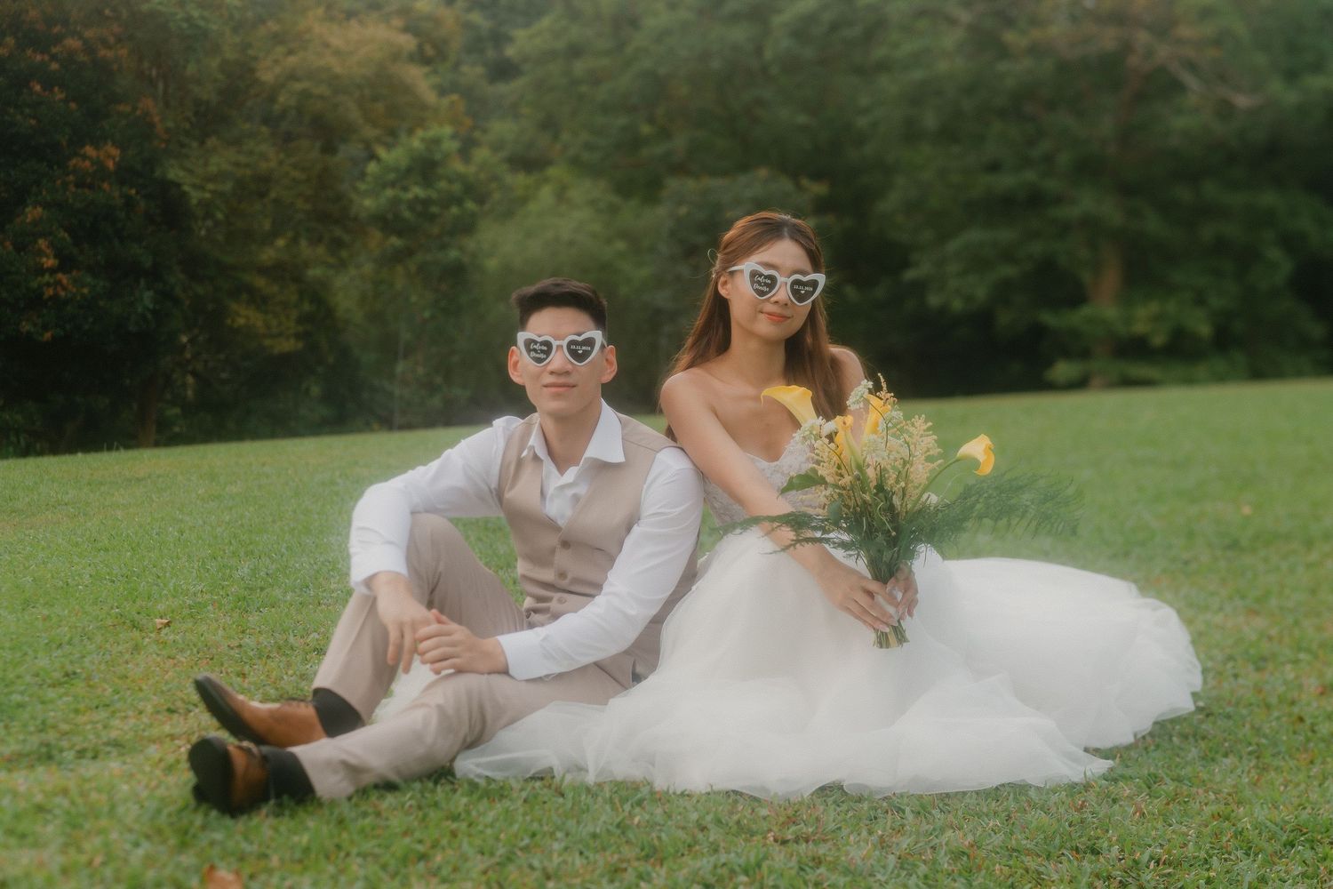 A playful wedding couple sitting on grass wearing sunglasses, with the bride in white and holding yellow flowers.