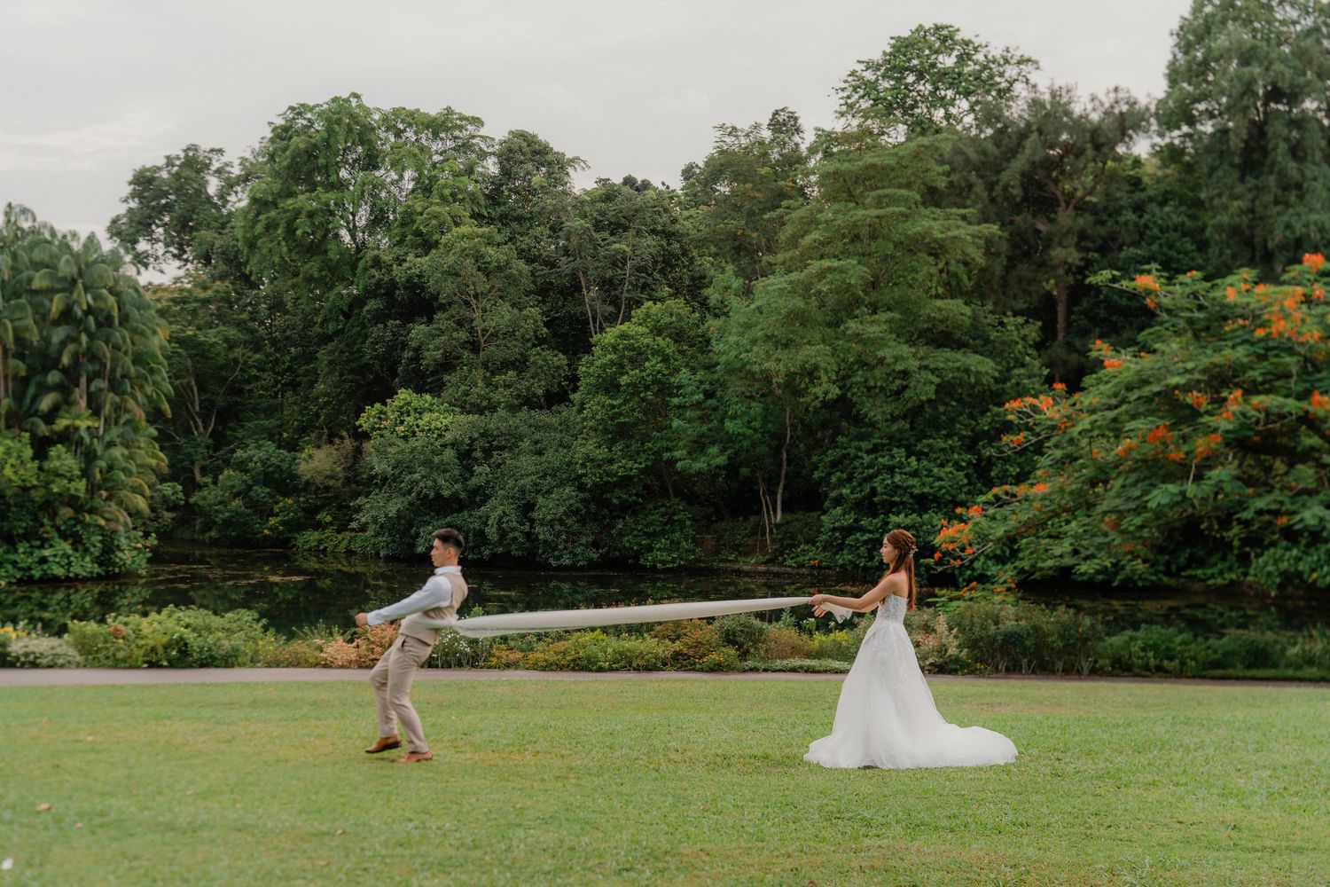 A couple plays a joyful game of catch on a lush green lawn surrounded by tall trees at an outdoor venue.