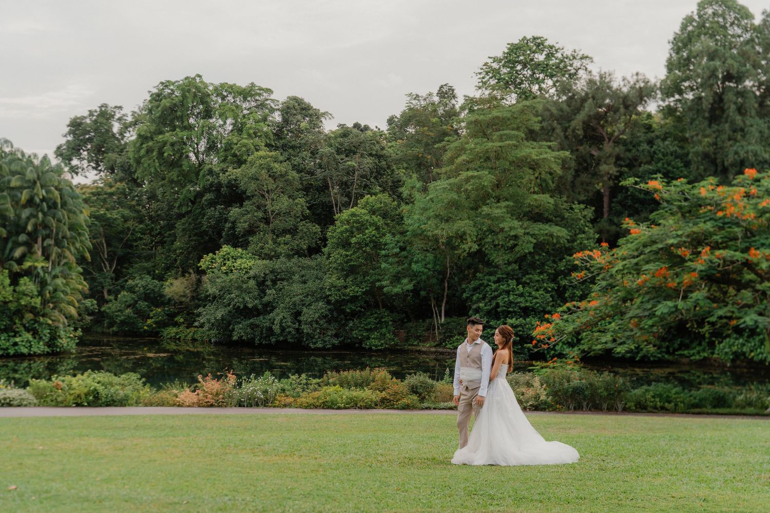 A couple stands together on a lush green lawn surrounded by tropical gardens and forest trees.