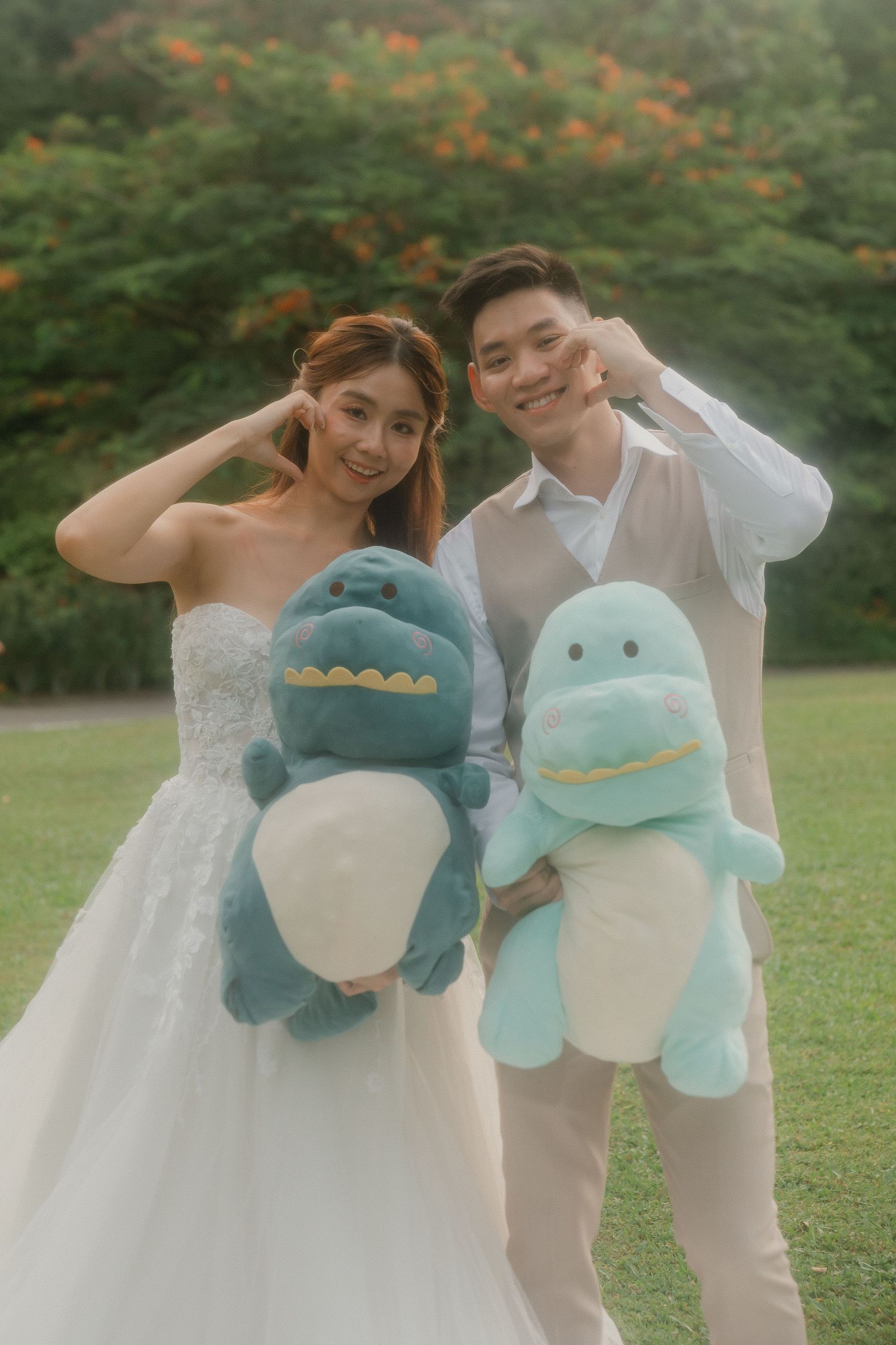 A couple in wedding attire poses outdoors holding matching blue and teal dinosaur plush toys on a grassy lawn.
