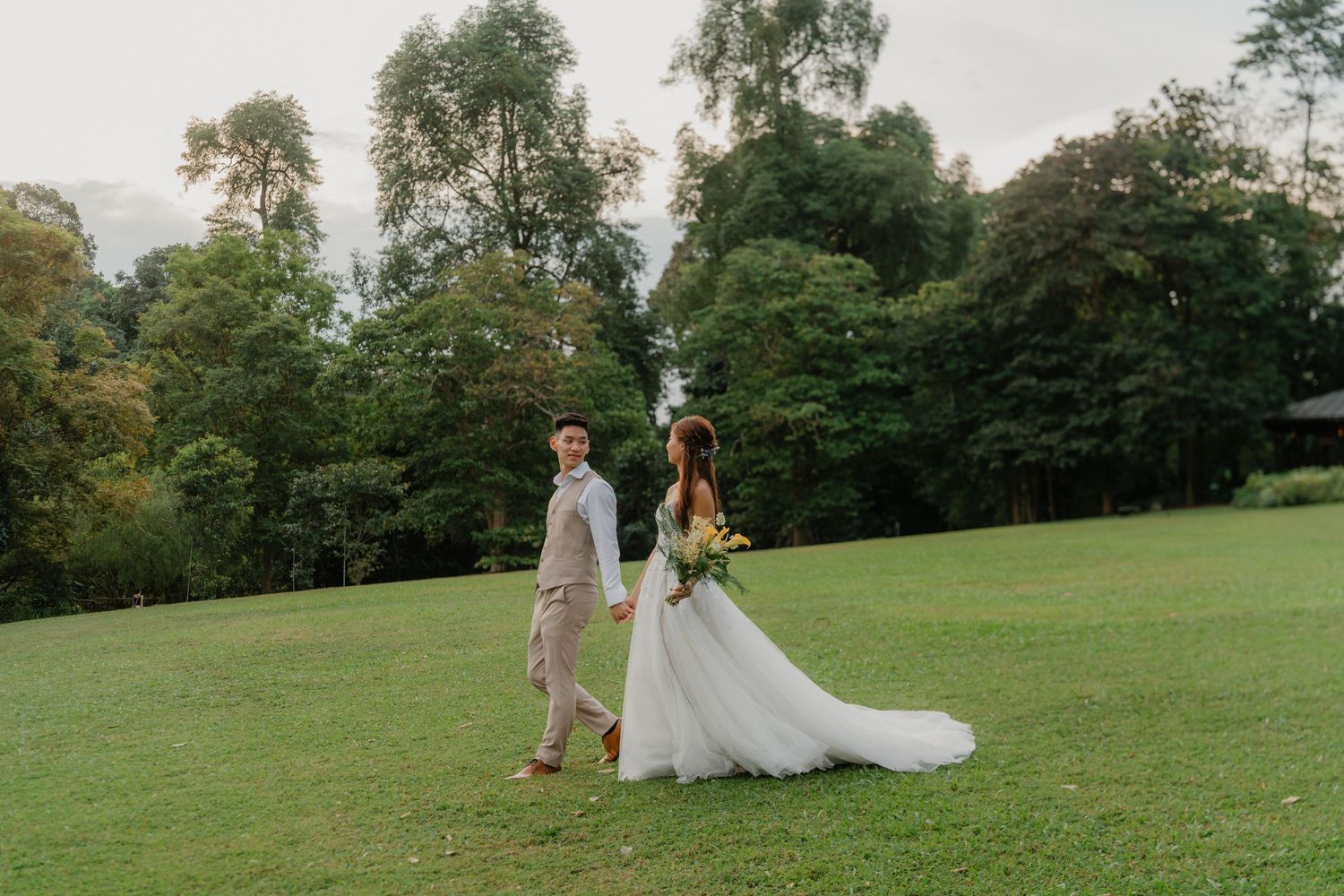 A wedding couple walks hand in hand across a serene hilltop meadow surrounded by trees on a sunny afternoon.