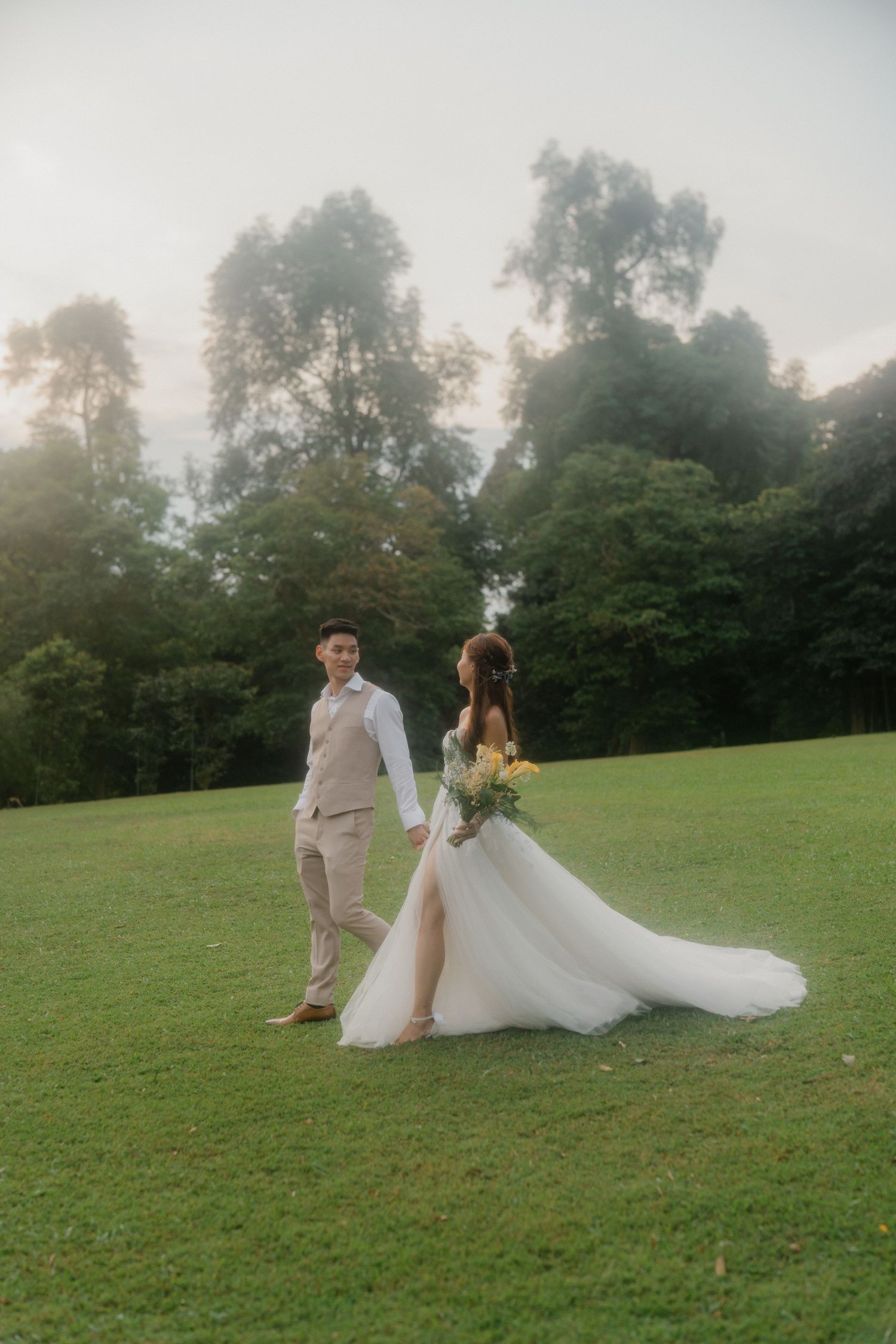 A couple in wedding attire poses for photographs on a misty grassy hill during golden hour.