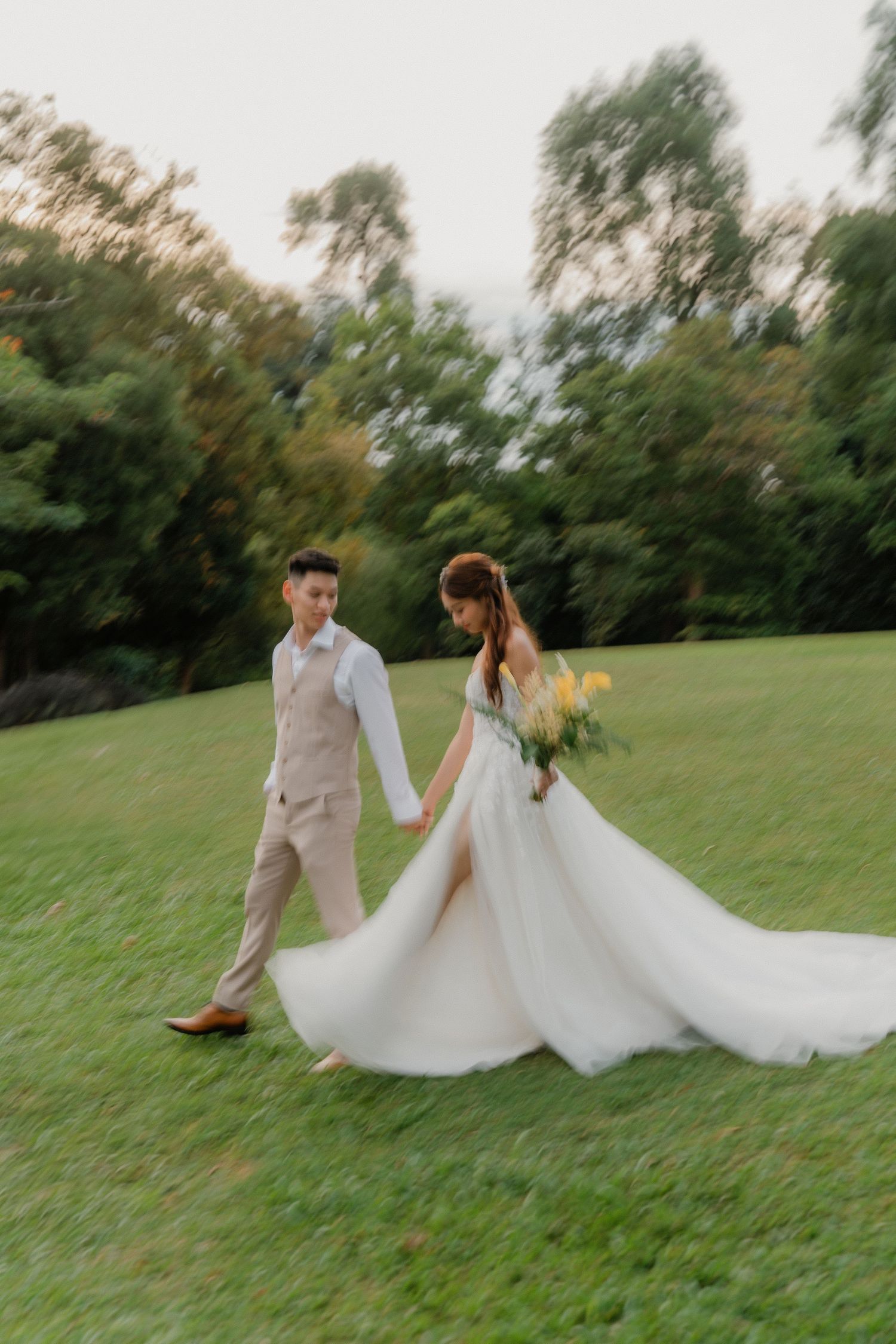 A couple in wedding attire twirls and dances together on a grassy hillside in a romantic outdoor setting.