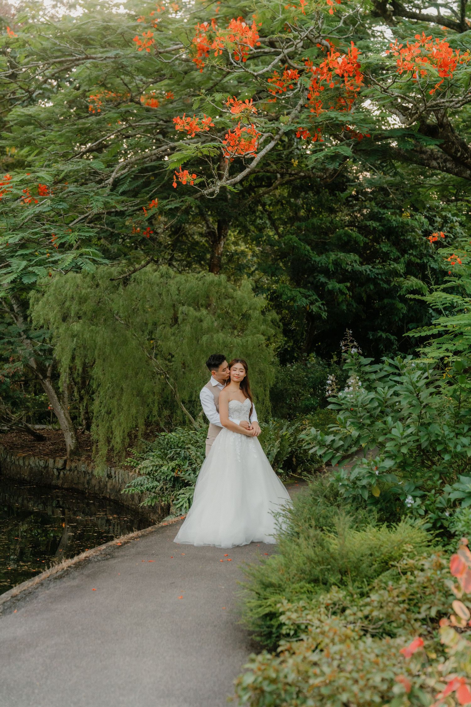 A couple poses under vibrant orange flowers and lush greenery along a garden path on their wedding day.