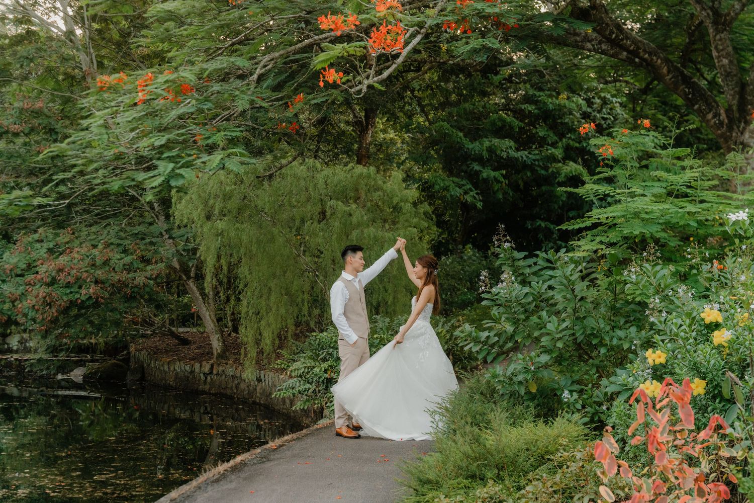 Bride in white wedding dress walking through lush forest garden path with orange wildflowers blooming nearby.