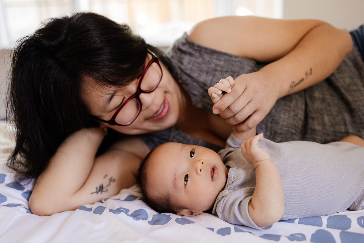 A mom lays on a bed with her newborn baby who holds her hand