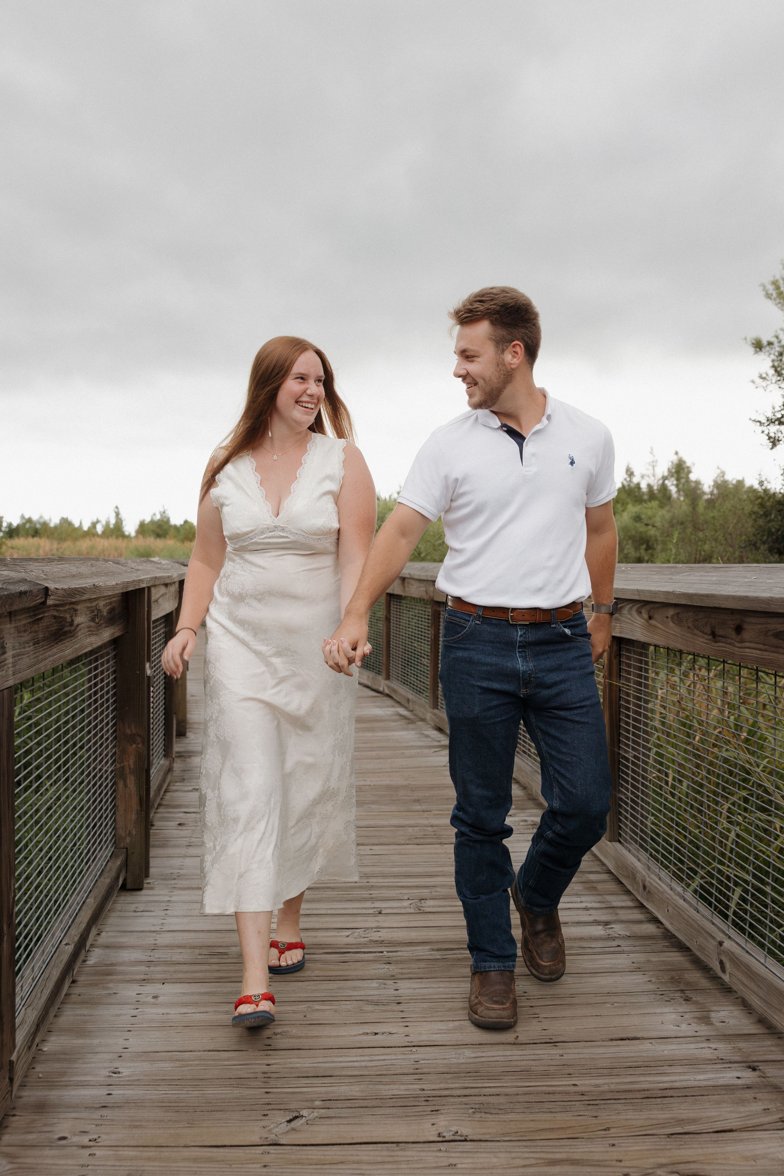 A couple walking and laughing together on a wooden boardwalk surrounded by greenery.