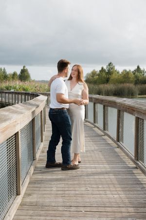 A couple in white and denim embraces on a wooden boardwalk over wetlands.