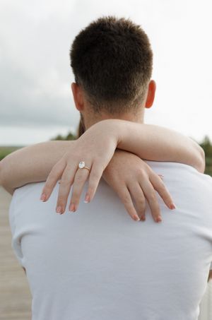Hands with an engagement ring embrace around shoulders in a white shirt.