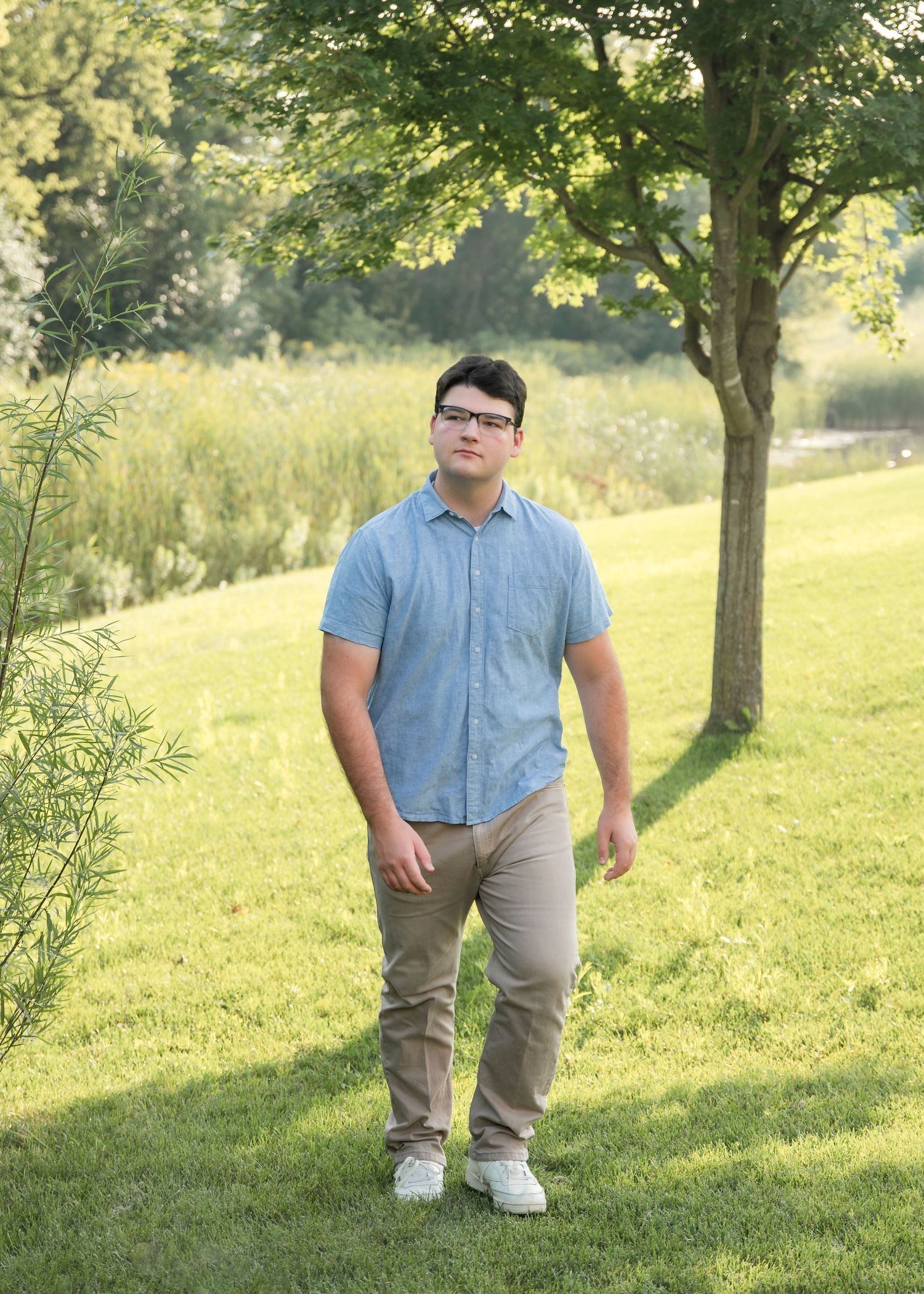 A person in casual attire stands on a grassy hillside beneath trees on a sunny day.