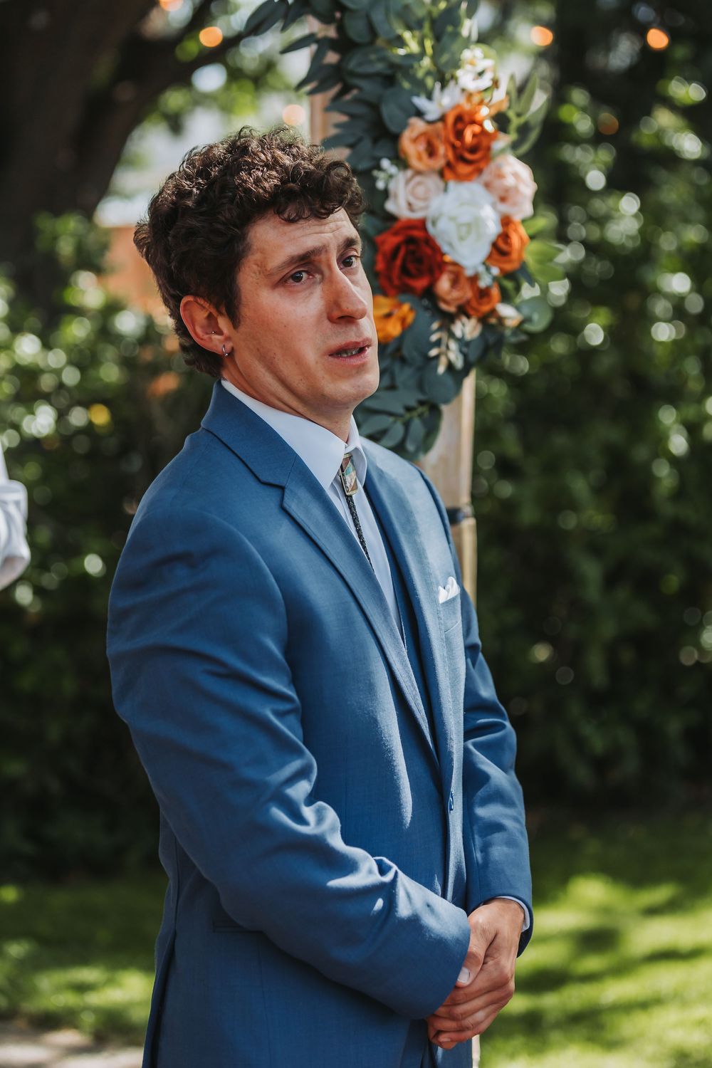 Groom in a navy blue suit stands outdoors during a wedding ceremony with floral decor and natural greenery in background.