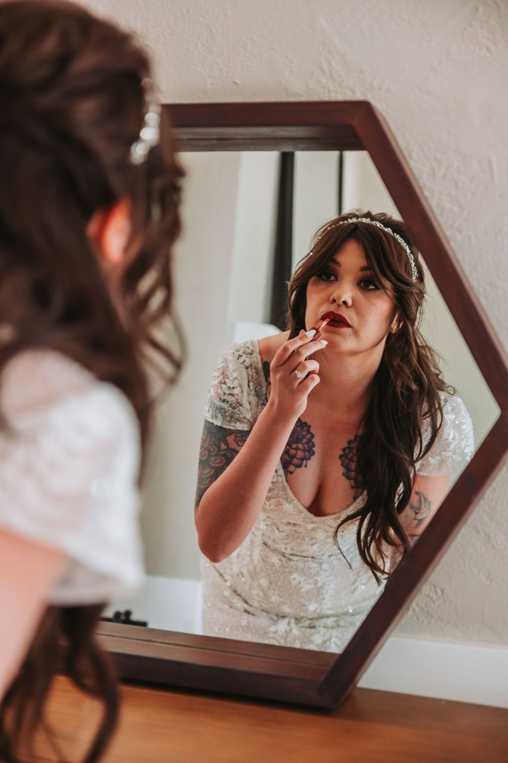 A reflection in a hexagonal mirror shows someone applying lipstick while wearing a delicate lace wedding dress.