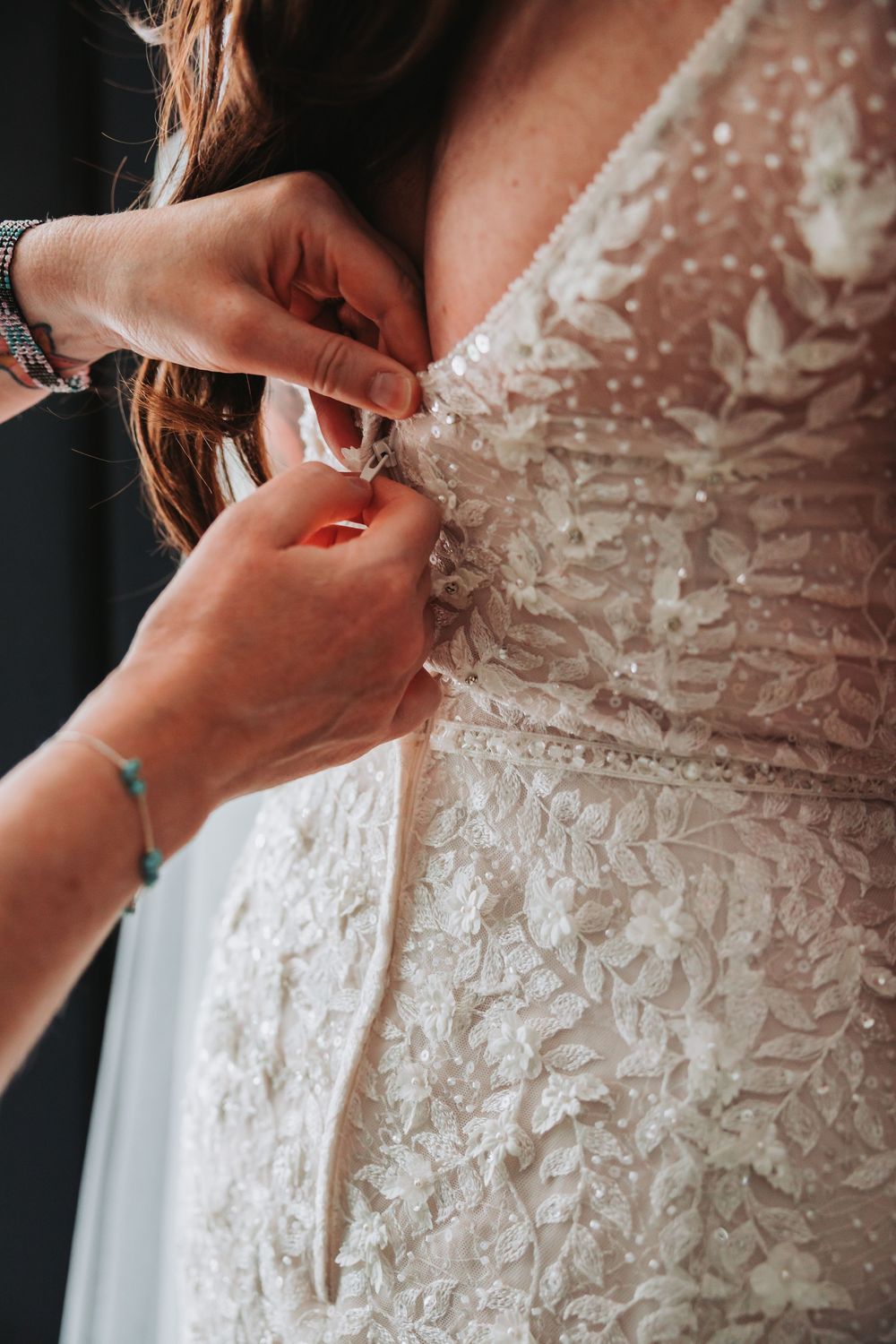 Close-up shots of hands fastening delicate lace and buttons on the back of an ornate white wedding dress.