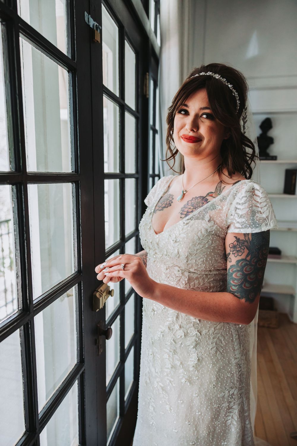A bride in a delicate lace wedding dress with short sleeves stands by a window in natural light, wearing a crystal headpiece.